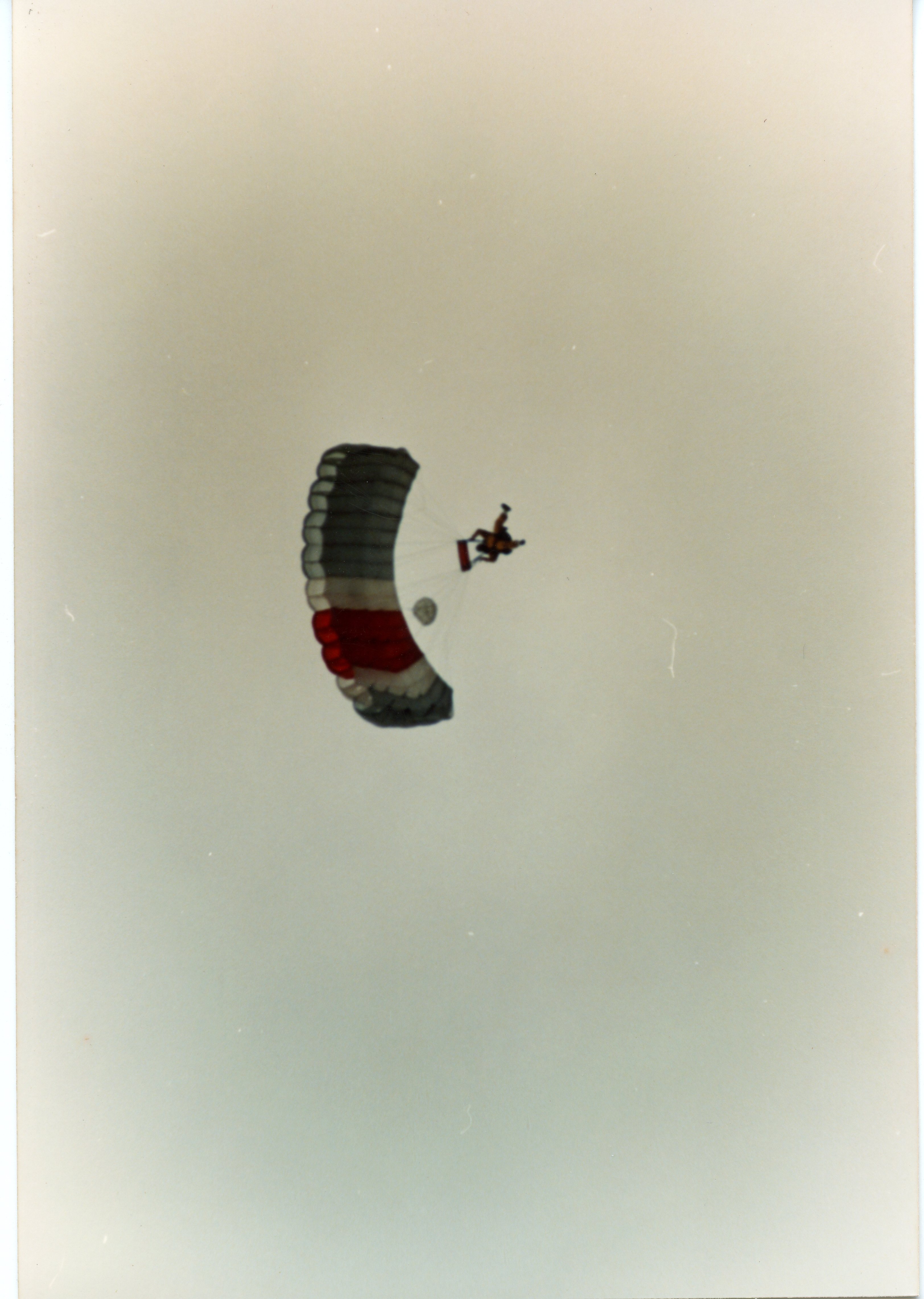 Uma fotografia aérea capturando um paraquedista em pleno voo, utilizando um paraquedas de formato retangular com faixas nas cores cinza, branco e vermelho. A imagem apresenta um céu limpo e sem nuvens, sugerindo uma atividade esportiva ou de treinamento realizada em um dia de boa visibilidade.