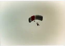 Uma fotografia aérea capturando um paraquedista em pleno voo, utilizando um paraquedas de formato retangular com faixas nas cores cinza, branco e vermelho. A imagem apresenta um céu limpo e sem nuvens, sugerindo uma atividade esportiva ou de treinamento realizada em um dia de boa visibilidade.