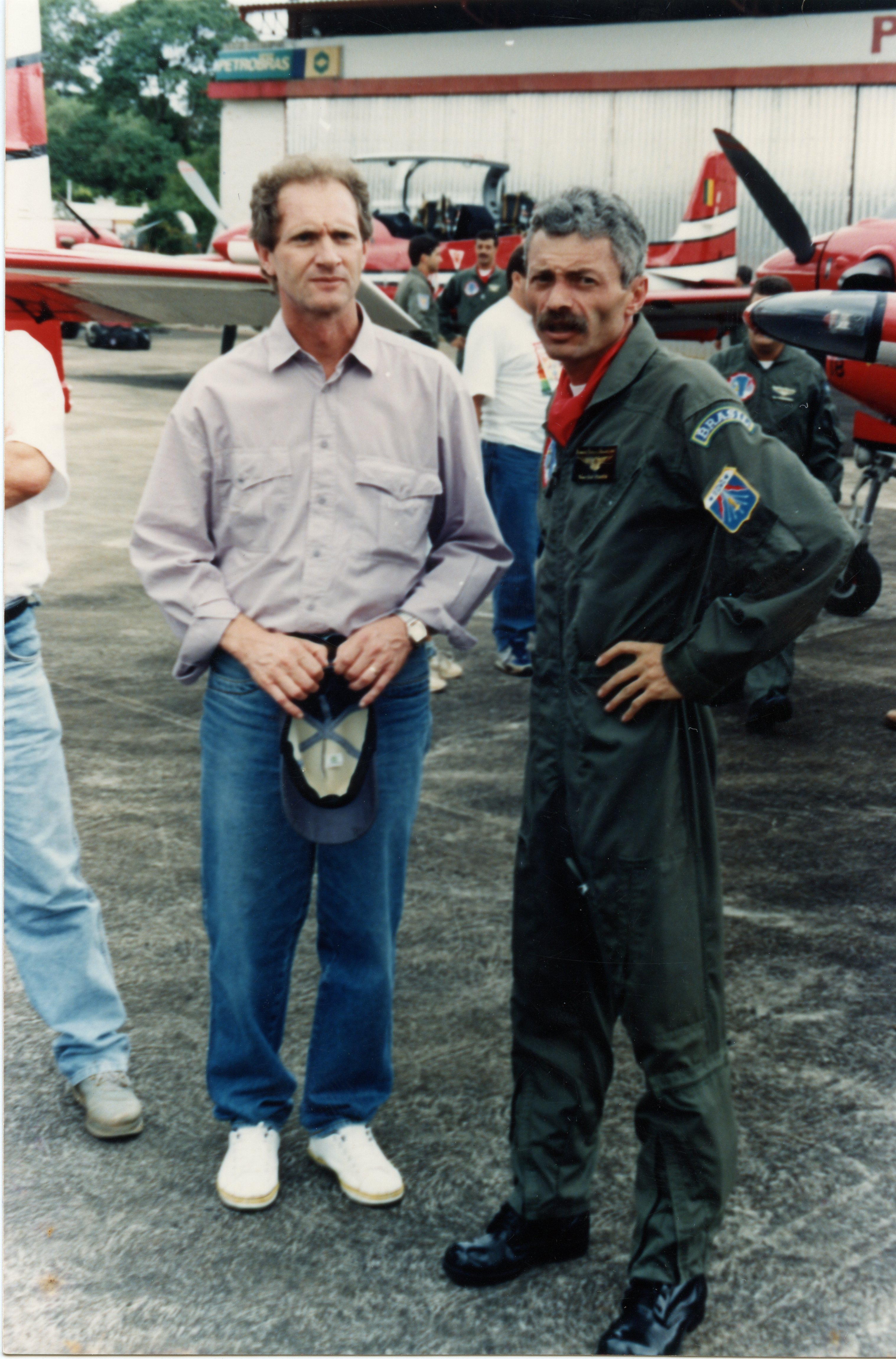Uma fotografia colorida mostrando dois homens em primeiro plano em um pátio de hangar de aeroporto, com aeronaves ao fundo. Um dos homens veste roupas casuais e o outro está uniformizado como piloto, sugerindo um contexto de aviação civil ou militar no Brasil, possivelmente nas décadas de 1980 ou 1990.