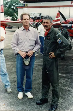 Uma fotografia colorida mostrando dois homens em primeiro plano em um pátio de hangar de aeroporto, com aeronaves ao fundo. Um dos homens veste roupas casuais e o outro está uniformizado como piloto, sugerindo um contexto de aviação civil ou militar no Brasil, possivelmente nas décadas de 1980 ou 1990.