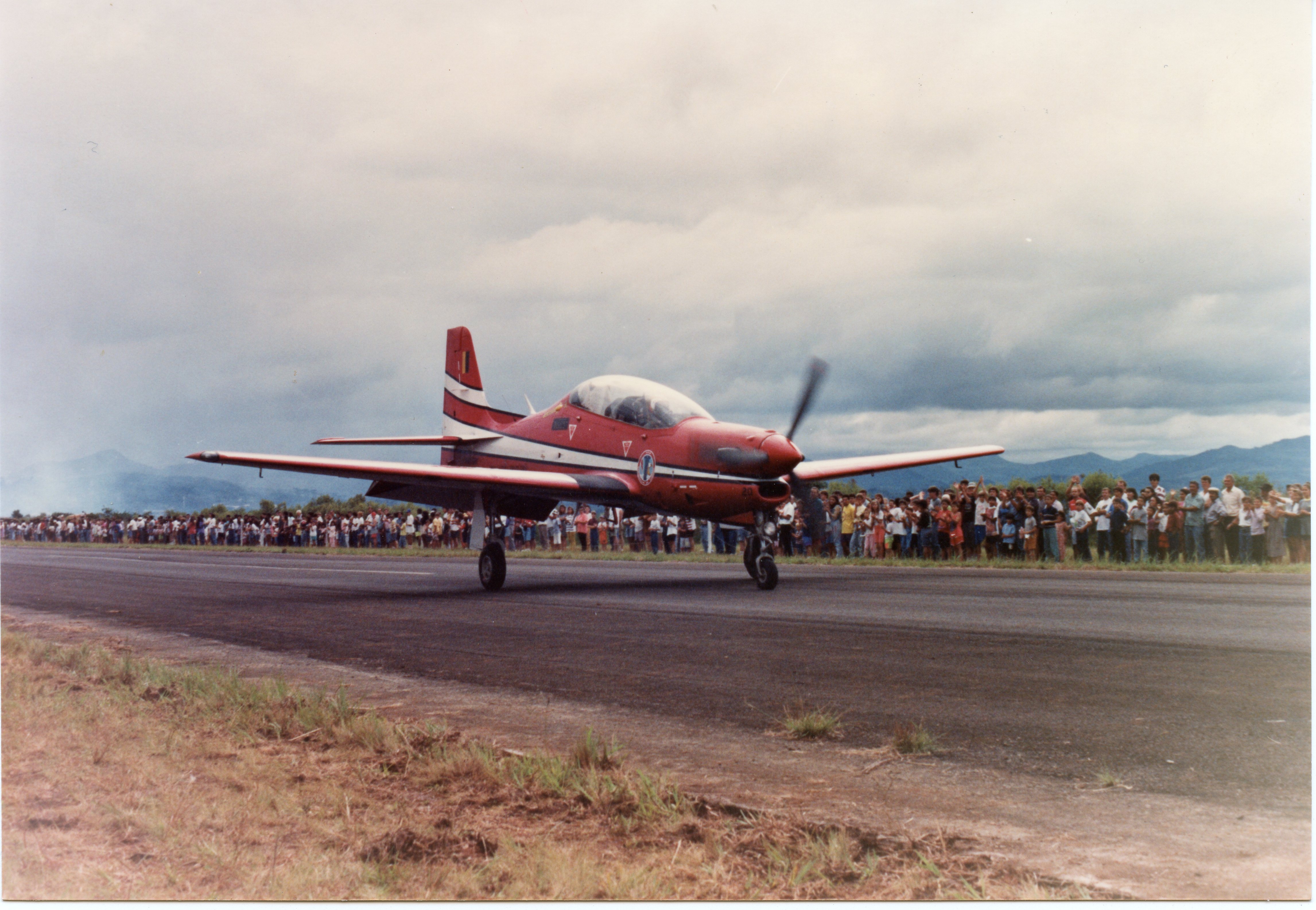 Uma fotografia colorida que mostra uma aeronave de treinamento militar, possivelmente um Embraer T-27 Tucano, taxiando em uma pista de pouso. Ao fundo, uma grande multidão de pessoas observa o evento, sugerindo uma demonstração aérea ou evento público em um aeródromo regional.