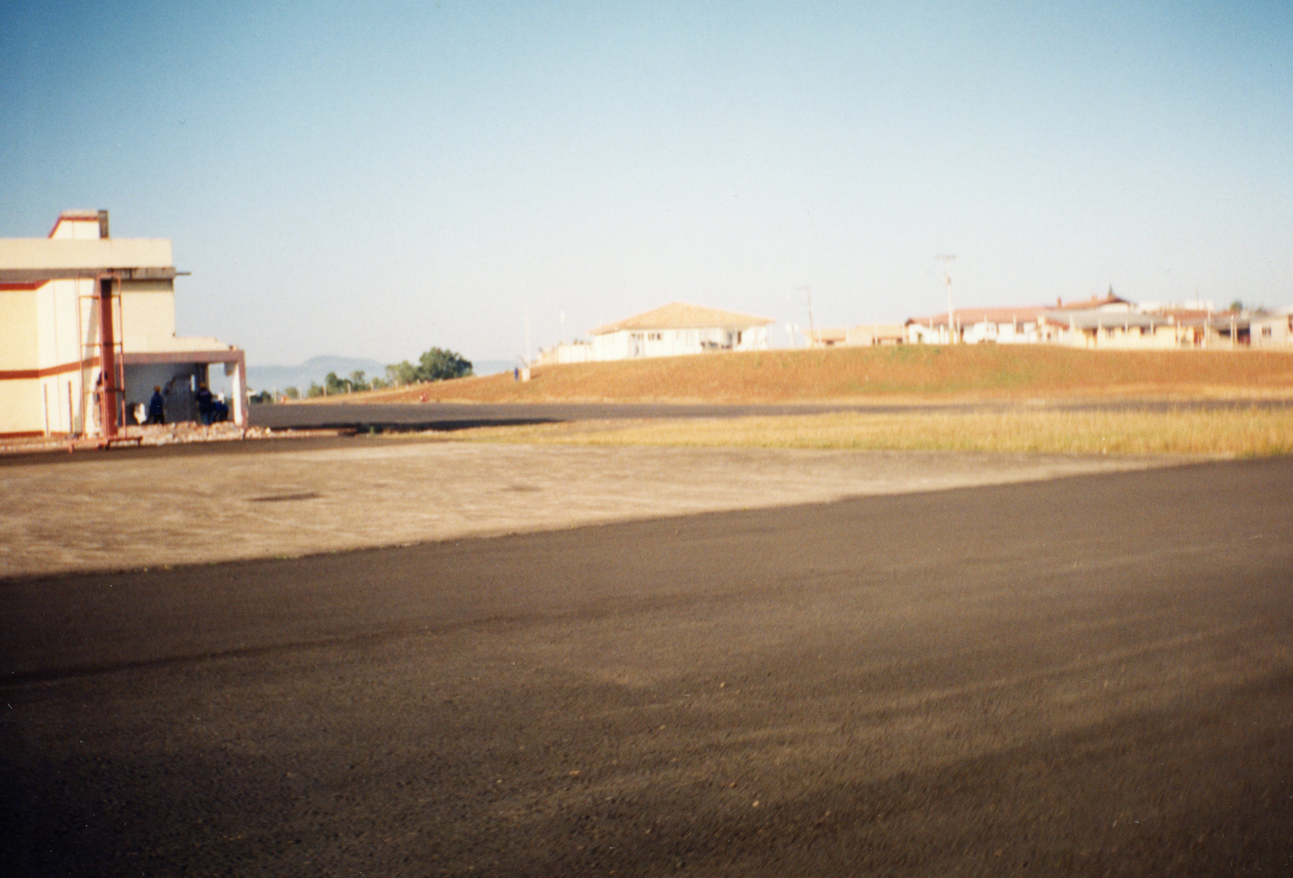 Uma fotografia de uma pista de pouso ou área pavimentada em uma região de topografia elevada, possivelmente em uma cidade do interior brasileiro. Ao fundo, observam-se construções residenciais simples e uma paisagem aberta, sugerindo um ambiente de aeródromo regional ou área industrial em expansão, provavelmente datada entre as décadas de 1980 e 1990.
