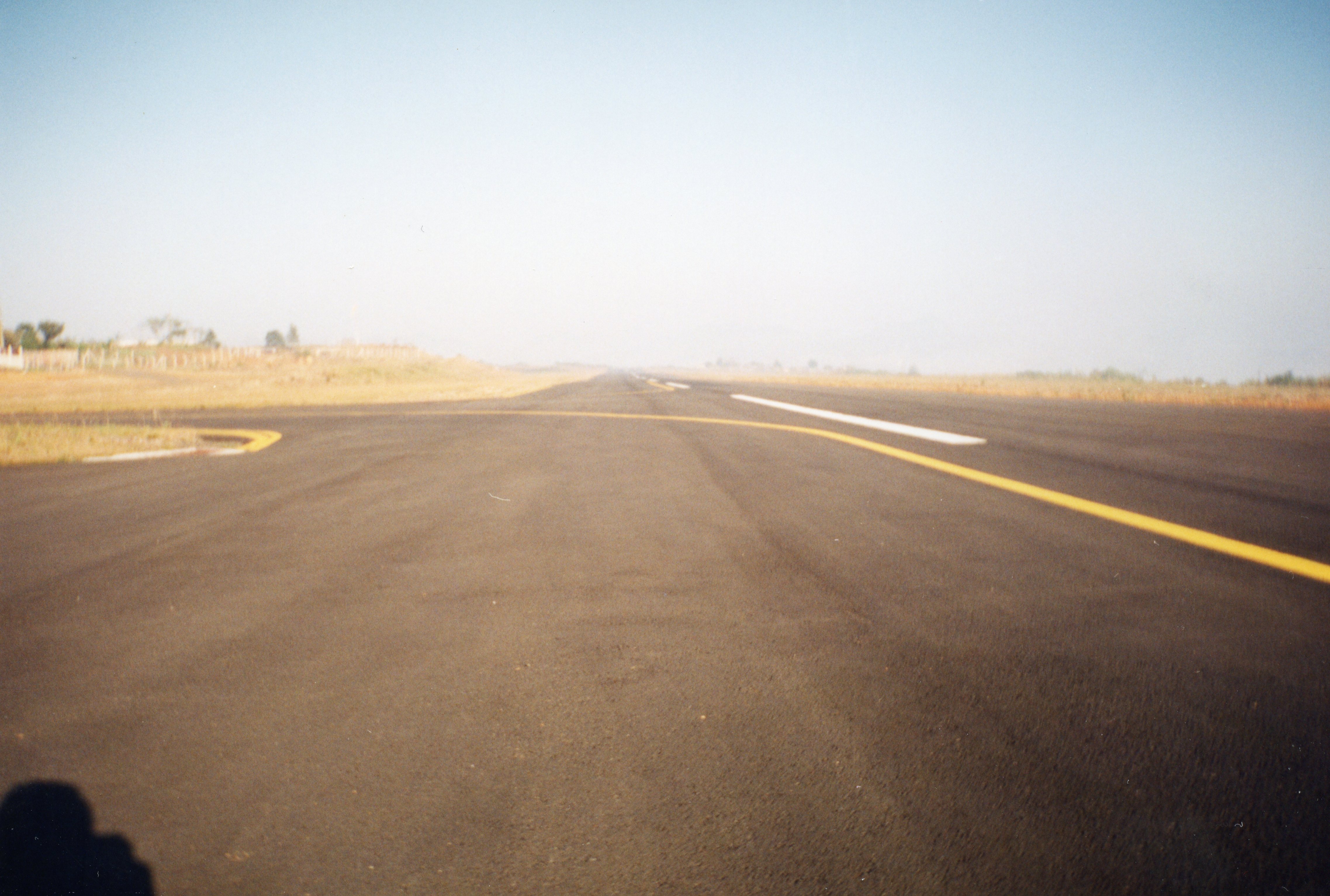 Uma fotografia de uma pista de pouso e decolagem asfaltada, possivelmente de um aeródromo regional de pequeno porte. A imagem apresenta uma perspectiva centralizada da pista com marcações de sinalização horizontal amarela e branca, sob um céu claro e horizonte levemente nebuloso.
