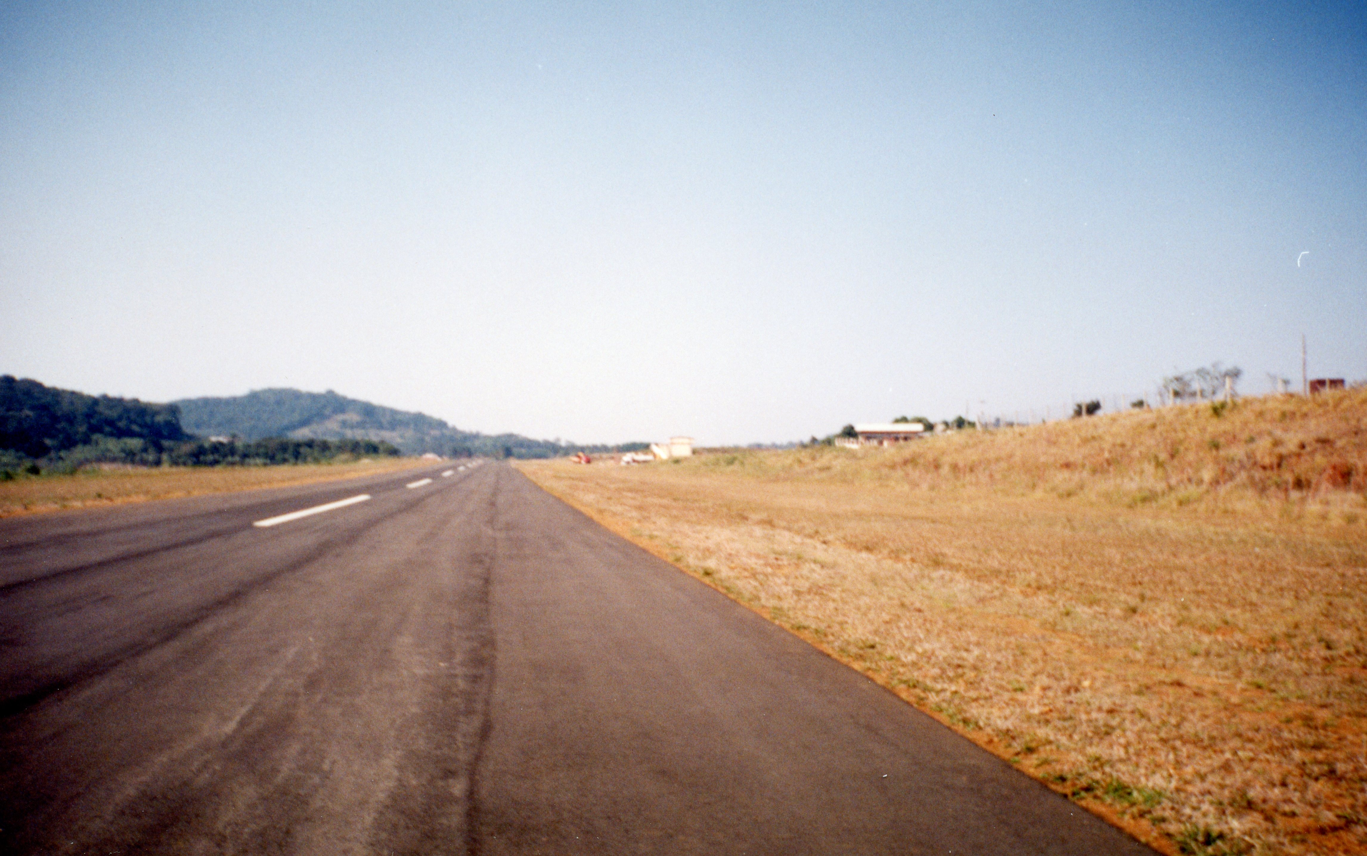 Uma fotografia de uma pista de pouso asfaltada, possivelmente de um aeródromo regional ou municipal, cercada por vegetação rasteira e colinas ao fundo. A imagem apresenta características de fotografia analógica do final do século XX, sugerindo uma infraestrutura de transporte em uma área rural ou de expansão urbana.