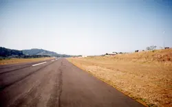 Uma fotografia de uma pista de pouso asfaltada, possivelmente de um aeródromo regional ou municipal, cercada por vegetação rasteira e colinas ao fundo. A imagem apresenta características de fotografia analógica do final do século XX, sugerindo uma infraestrutura de transporte em uma área rural ou de expansão urbana.