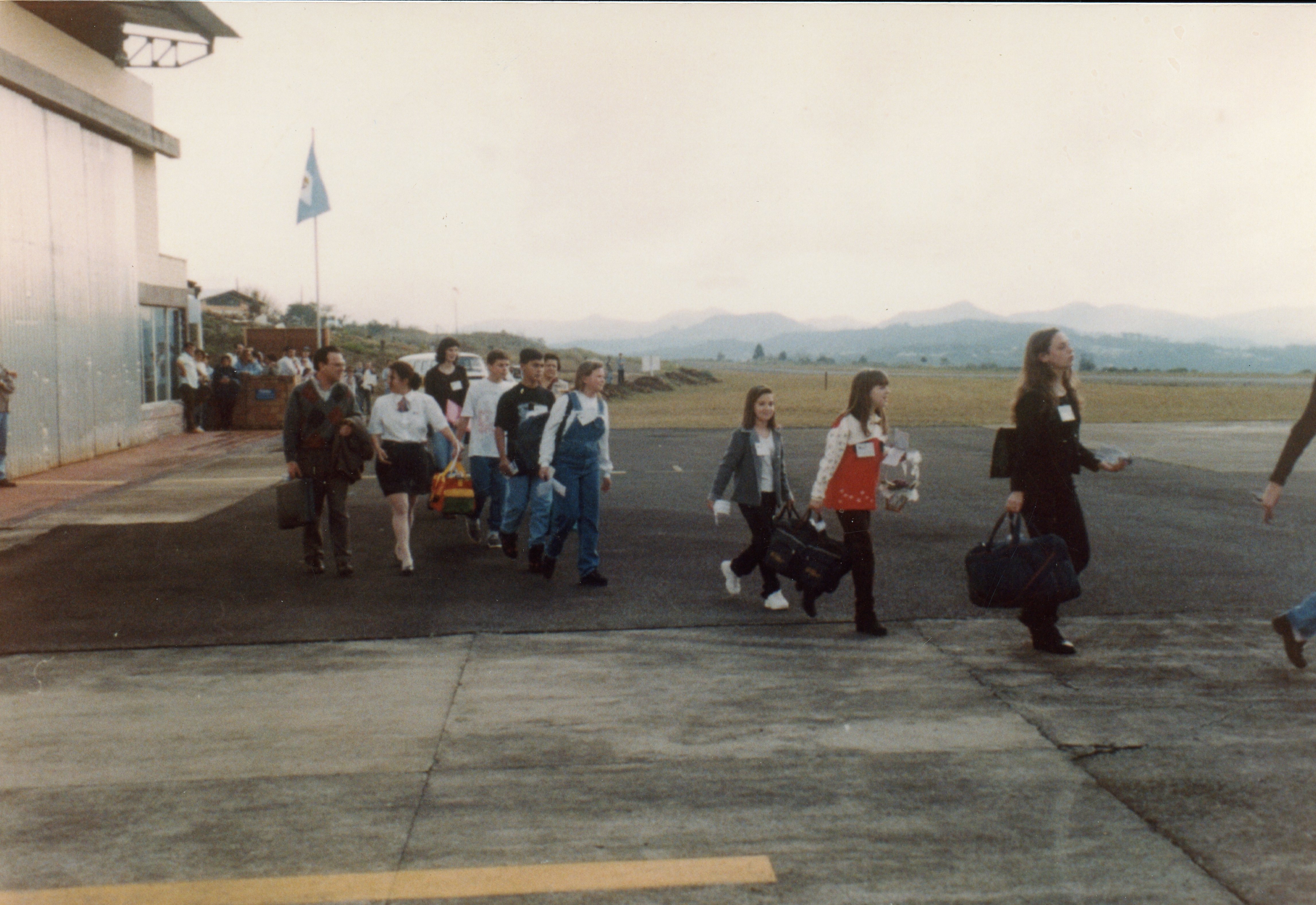 Uma fotografia colorida que mostra um grupo de pessoas, incluindo jovens e adultos, caminhando em um pátio de aeroporto regional. As vestimentas e o estilo das malas sugerem um contexto de viagem escolar ou excursão ocorrida provavelmente na década de 1990.