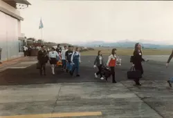 Uma fotografia colorida que mostra um grupo de pessoas, incluindo jovens e adultos, caminhando em um pátio de aeroporto regional. As vestimentas e o estilo das malas sugerem um contexto de viagem escolar ou excursão ocorrida provavelmente na década de 1990.