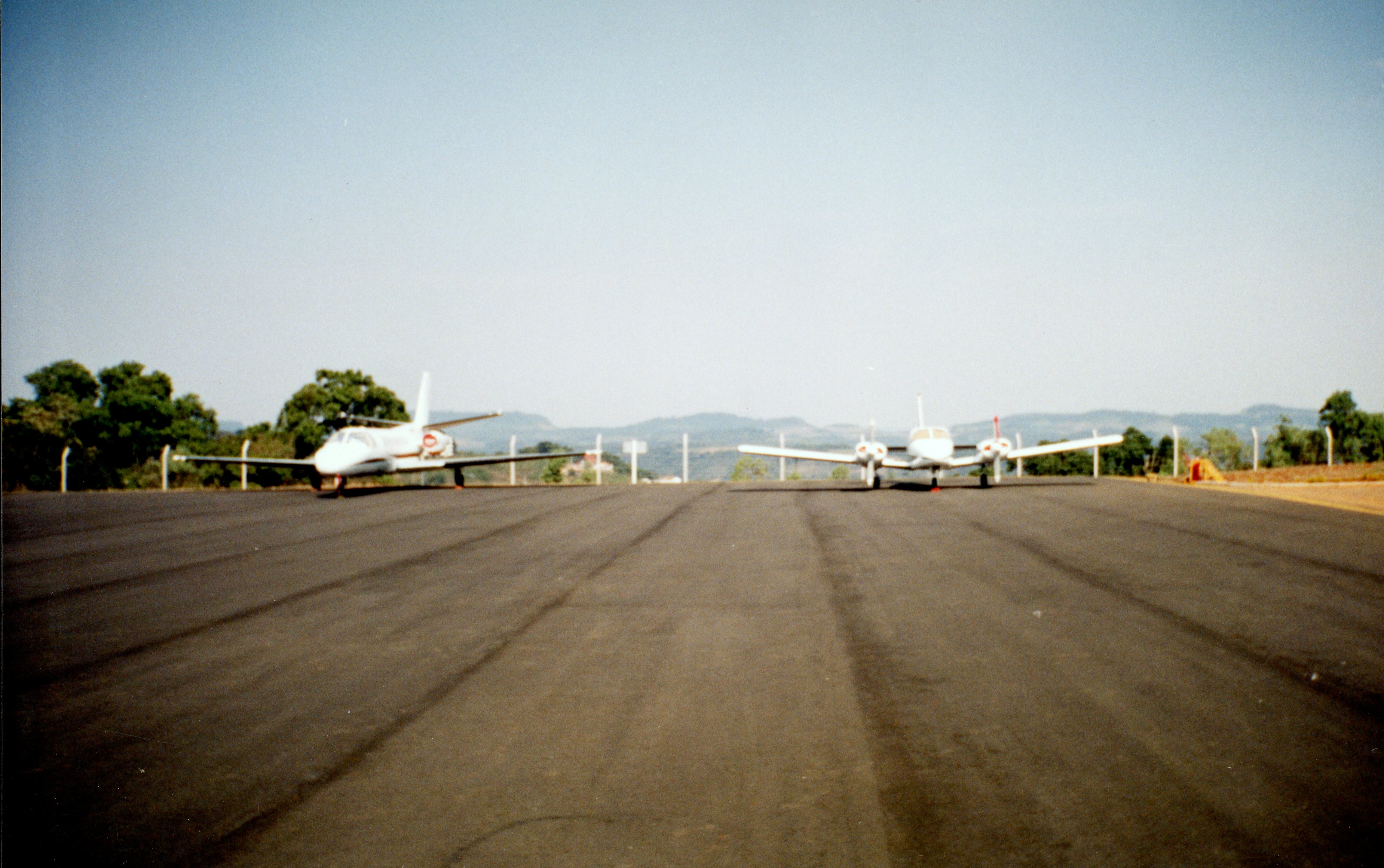 Uma fotografia colorida mostrando duas aeronaves estacionadas em uma pista de pouso asfaltada, possivelmente em um aeroporto regional brasileiro. O cenário apresenta um horizonte com colinas ao fundo e uma cerca de proteção, sugerindo uma infraestrutura de aviação de pequeno ou médio porte.