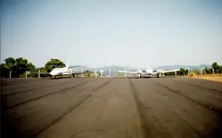 Uma fotografia colorida mostrando duas aeronaves estacionadas em uma pista de pouso asfaltada, possivelmente em um aeroporto regional brasileiro. O cenário apresenta um horizonte com colinas ao fundo e uma cerca de proteção, sugerindo uma infraestrutura de aviação de pequeno ou médio porte.