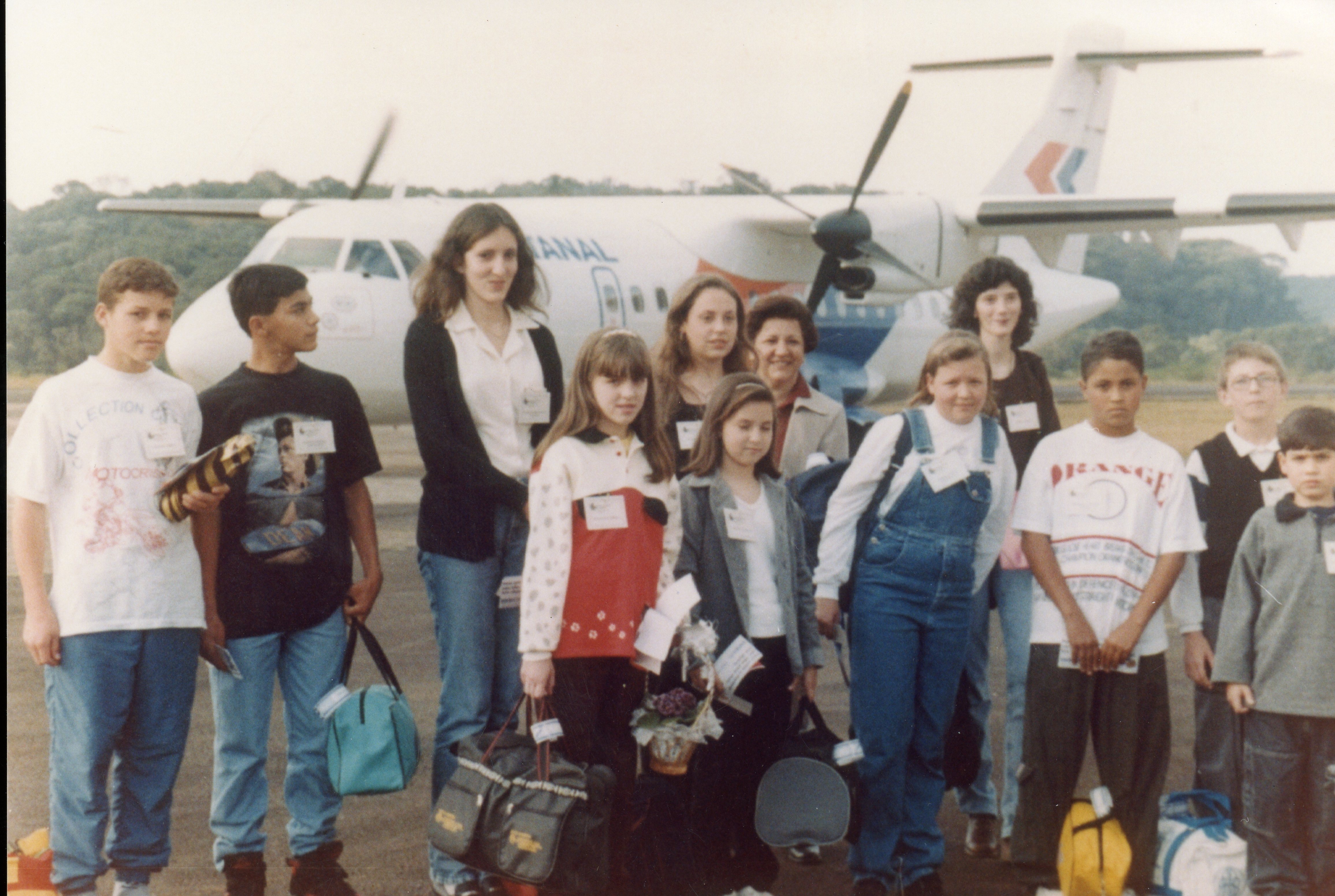 Uma fotografia de grupo de jovens e adultos posando em uma pista de aeroporto, com uma aeronave da companhia aérea Rio Sul ao fundo. A imagem sugere uma excursão escolar ou evento de visitação, com os participantes usando crachás de identificação e vestimentas típicas da década de 1990.