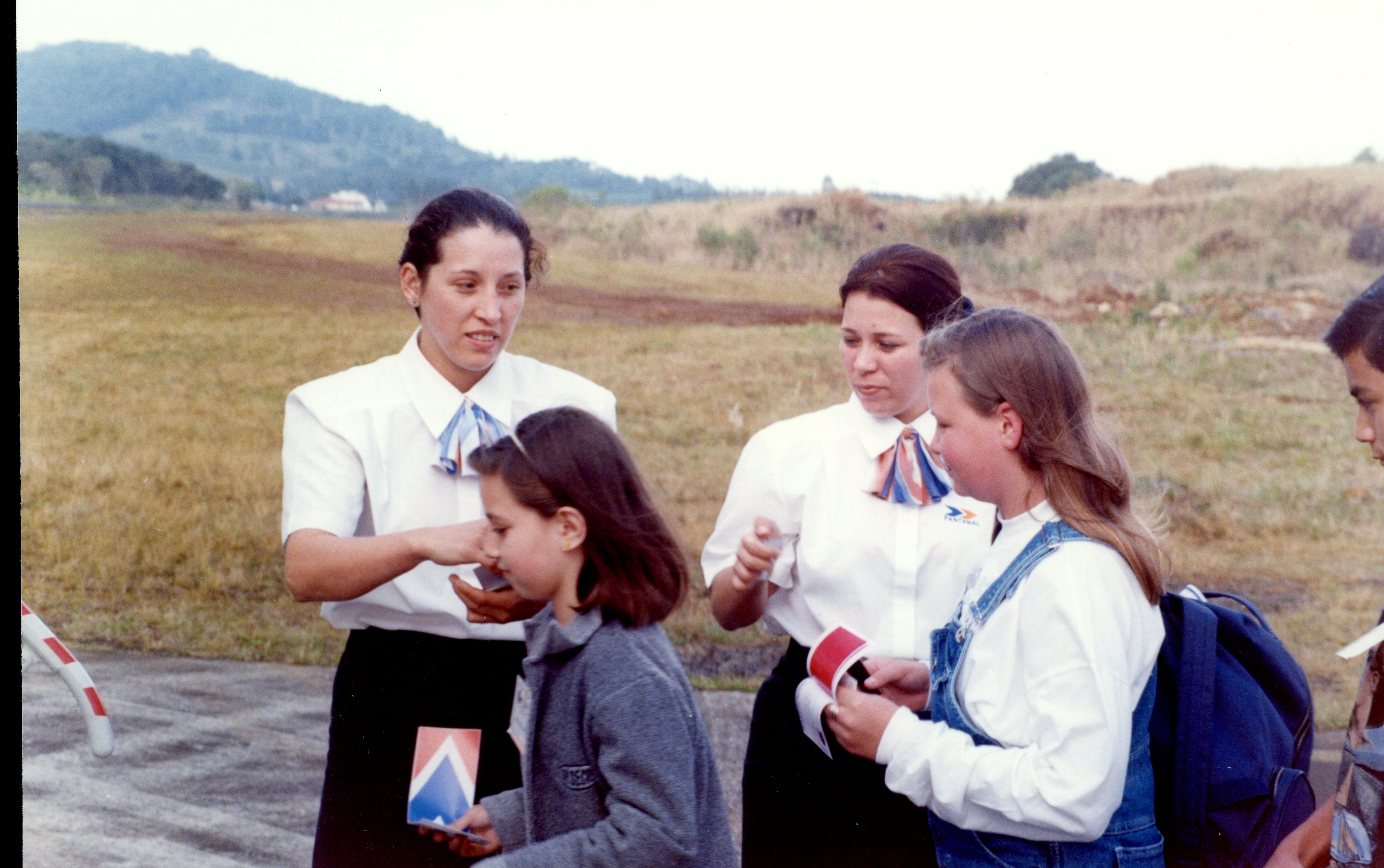 Uma fotografia colorida que mostra duas mulheres vestindo uniformes corporativos, possivelmente funcionárias de uma companhia aérea ou evento, interagindo com duas jovens em uma área externa aberta. O cenário sugere um ambiente de aeroporto ou campo de aviação, com vegetação rasteira e colinas ao fundo, típico de registros fotográficos da década de 1990.