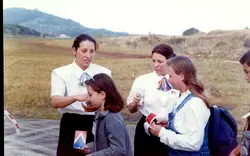 Uma fotografia colorida que mostra duas mulheres vestindo uniformes corporativos, possivelmente funcionárias de uma companhia aérea ou evento, interagindo com duas jovens em uma área externa aberta. O cenário sugere um ambiente de aeroporto ou campo de aviação, com vegetação rasteira e colinas ao fundo, típico de registros fotográficos da década de 1990.
