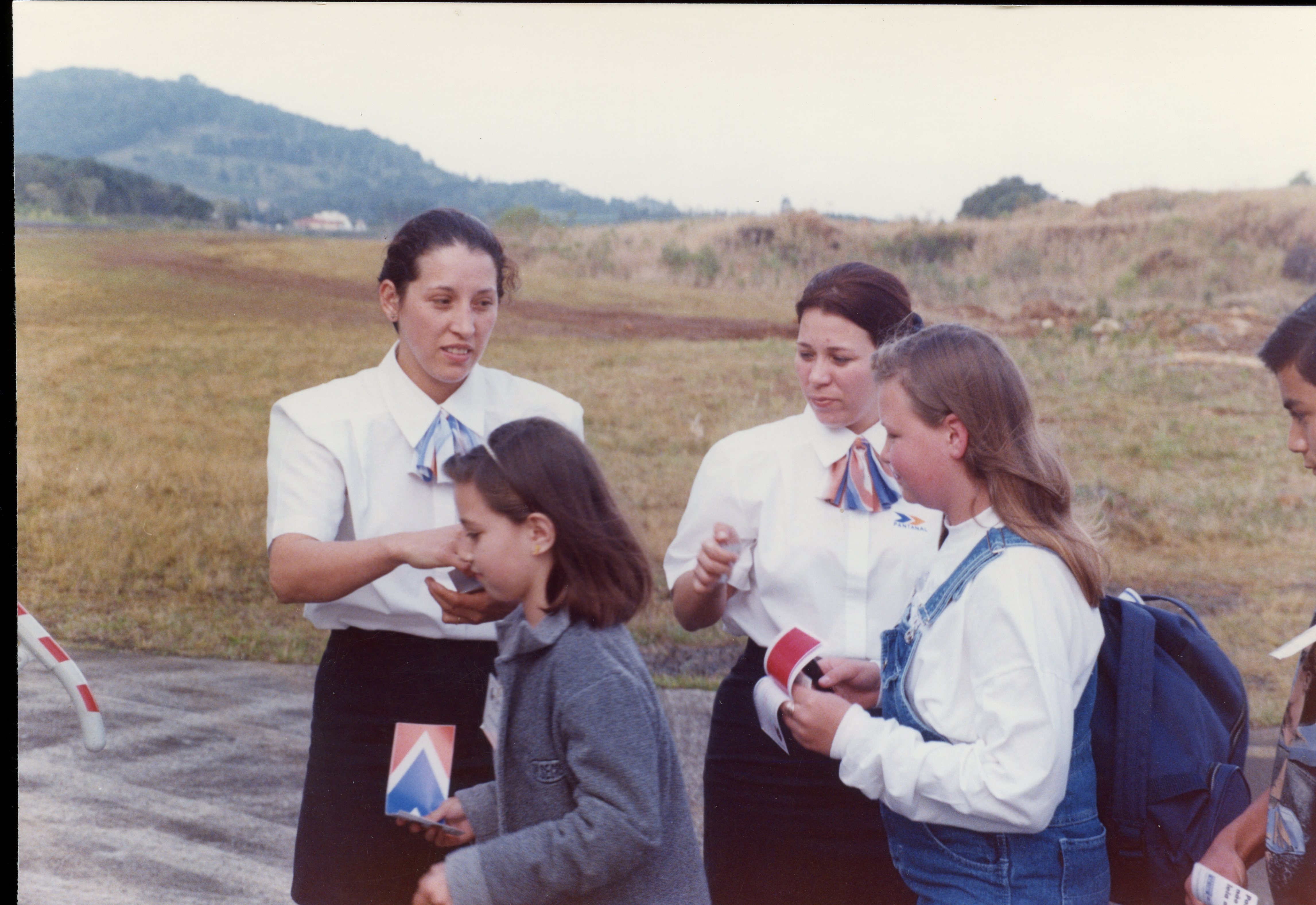 Uma fotografia colorida que mostra duas mulheres vestindo uniformes de comissárias de bordo da companhia aérea Pantanal, interagindo com crianças em uma área externa próxima a uma pista de pouso. A cena sugere um evento promocional ou uma visita guiada em um aeroporto regional, com vestimentas e estilo fotográfico típicos da década de 1990.