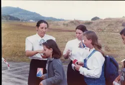 Uma fotografia colorida que mostra duas mulheres vestindo uniformes de comissárias de bordo da companhia aérea Pantanal, interagindo com crianças em uma área externa próxima a uma pista de pouso. A cena sugere um evento promocional ou uma visita guiada em um aeroporto regional, com vestimentas e estilo fotográfico típicos da década de 1990.