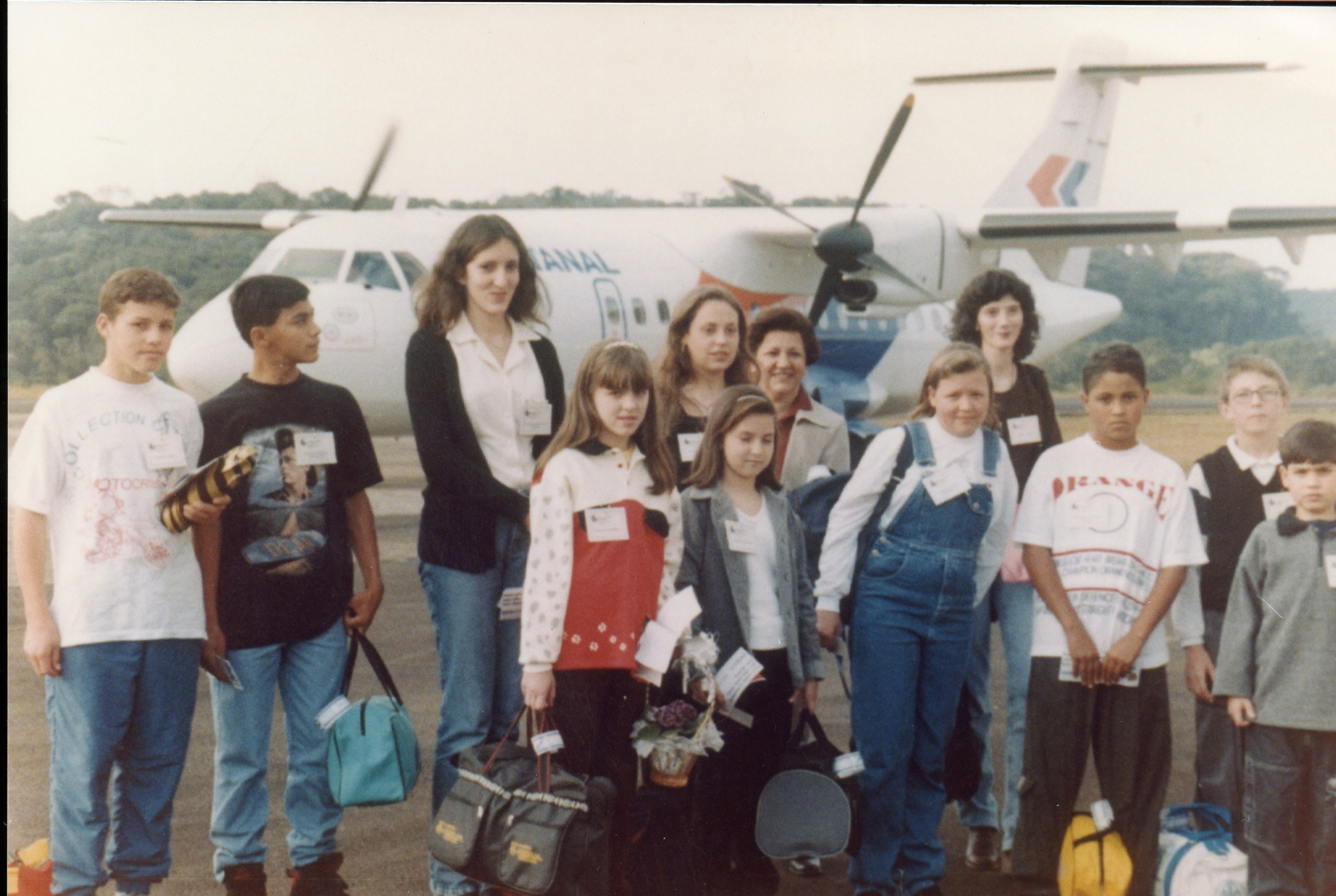 Uma fotografia de grupo de jovens e adultos posando em uma pista de aeroporto, com uma aeronave da companhia aérea Rio Sul ao fundo. A imagem sugere uma excursão escolar ou evento de visita técnica, com os participantes usando crachás e carregando bolsas, típica da moda e estilo de vida do final da década de 1990.