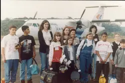 Uma fotografia de grupo de jovens e adultos posando em uma pista de aeroporto, com uma aeronave da companhia aérea Rio Sul ao fundo. A imagem sugere uma excursão escolar ou evento de visita técnica, com os participantes usando crachás e carregando bolsas, típica da moda e estilo de vida do final da década de 1990.