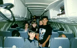 Uma fotografia interna de uma aeronave comercial mostrando um grupo de crianças e adolescentes sentados e em pé no corredor. O estilo das roupas e a qualidade da imagem sugerem um registro do final da década de 1990 ou início dos anos 2000, possivelmente durante uma excursão escolar ou viagem em grupo.