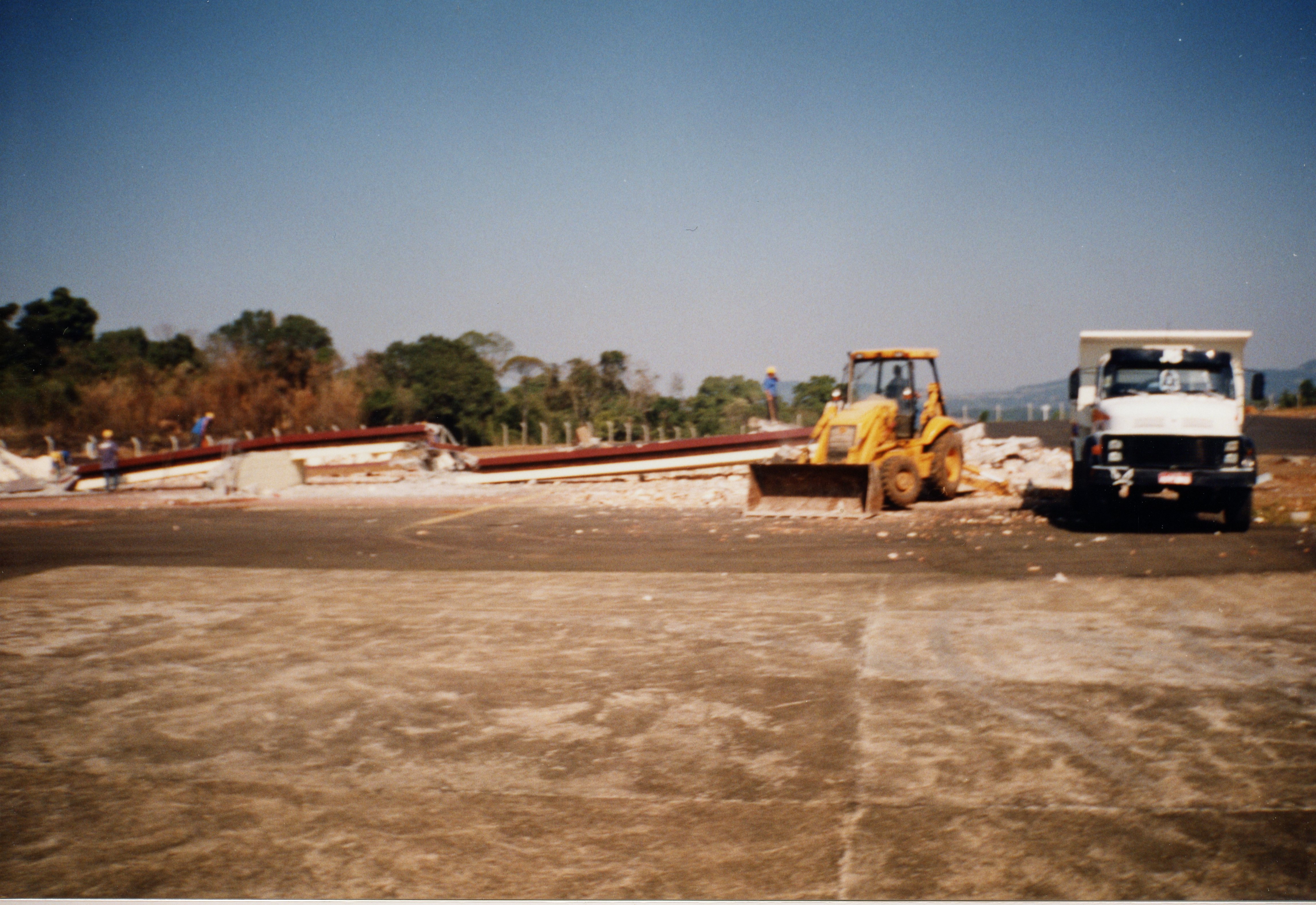 A imagem registra uma cena de demolição ou obra de infraestrutura em uma área externa, possivelmente um aeroporto ou pátio industrial. Observa-se uma retroescavadeira amarela e um caminhão basculante estacionados próximos a escombros de uma estrutura metálica, com trabalhadores presentes no local.