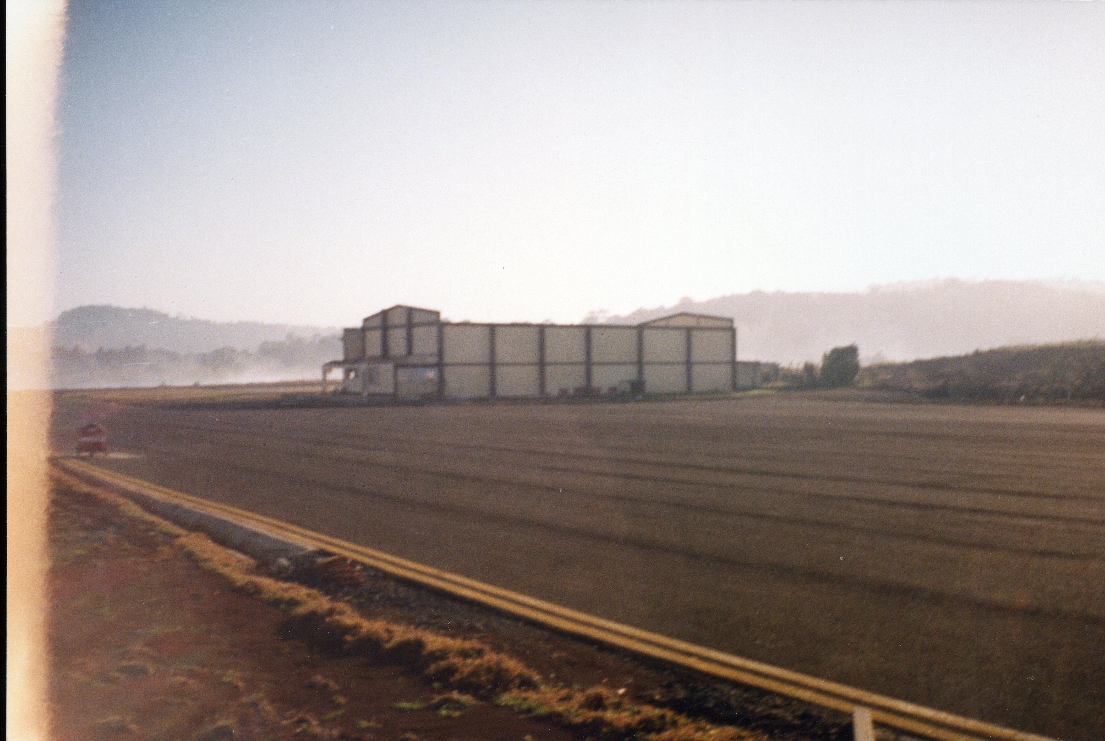 Uma fotografia de uma pista de pouso ou área industrial com um grande galpão ao fundo, sob uma luz matinal ou de fim de tarde com neblina. A imagem apresenta características de fotografia analógica amadora, possivelmente capturada em uma região rural ou de expansão industrial no Brasil.