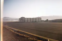 Uma fotografia de uma pista de pouso ou área industrial com um grande galpão ao fundo, sob uma luz matinal ou de fim de tarde com neblina. A imagem apresenta características de fotografia analógica amadora, possivelmente capturada em uma região rural ou de expansão industrial no Brasil.