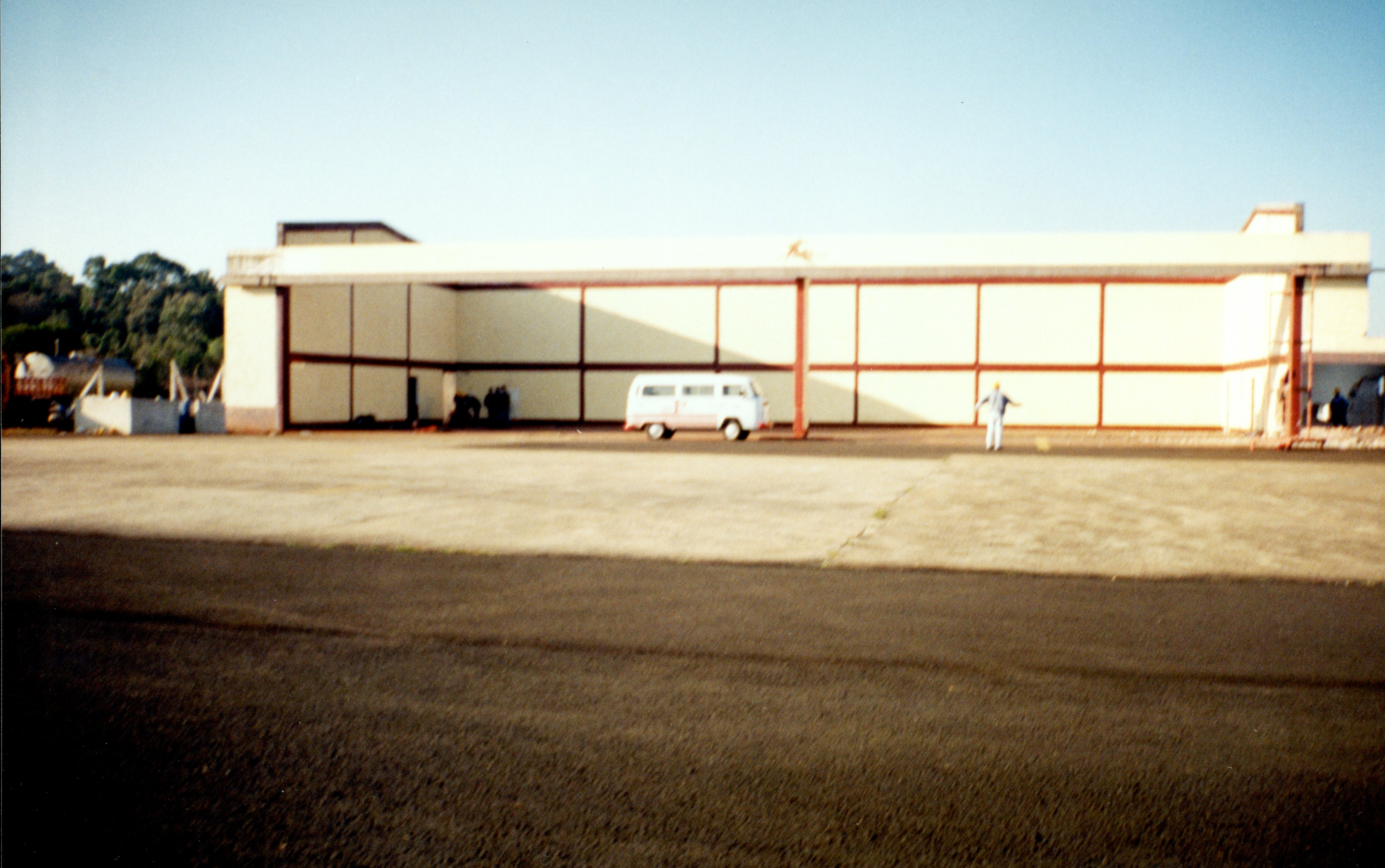 Uma fotografia de um grande hangar de aeroporto ou instalação industrial, possivelmente em uma cidade do interior brasileiro. A imagem mostra uma van Volkswagen Kombi estacionada em frente à estrutura, com uma pessoa caminhando próxima, sugerindo uma cena cotidiana de operação ou manutenção em meados da década de 1980 ou 1990.