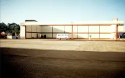 Uma fotografia de um grande hangar de aeroporto ou instalação industrial, possivelmente em uma cidade do interior brasileiro. A imagem mostra uma van Volkswagen Kombi estacionada em frente à estrutura, com uma pessoa caminhando próxima, sugerindo uma cena cotidiana de operação ou manutenção em meados da década de 1980 ou 1990.