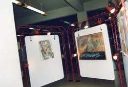 Uma exposição de arte contemporânea montada em um ambiente interno, utilizando estruturas metálicas modulares vermelhas para sustentar painéis brancos com pinturas abstratas. A iluminação é feita por refletores direcionados, sugerindo um evento cultural ou galeria de arte, provavelmente datado entre o final do século XX e início do XXI.