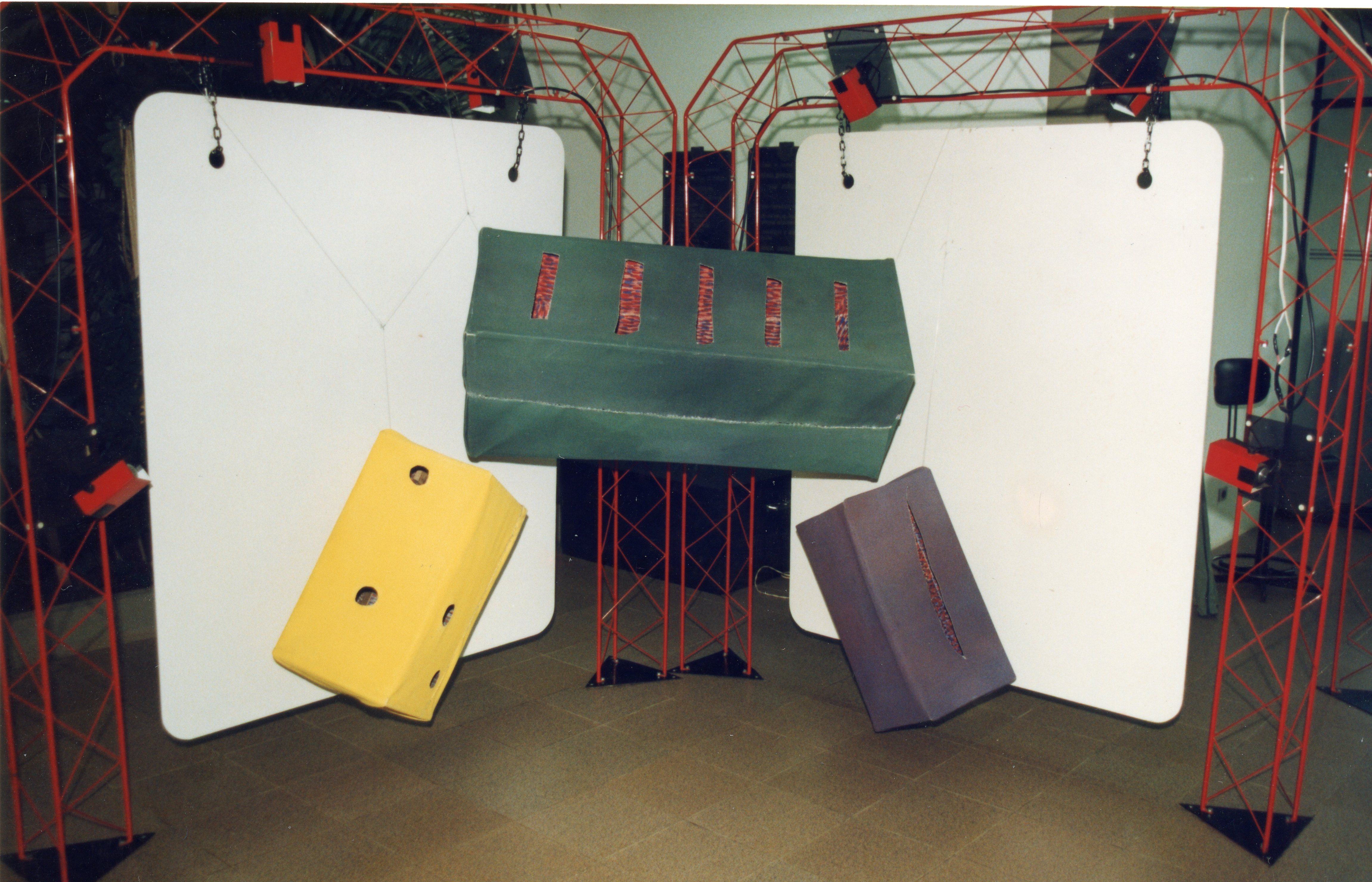 Uma instalação artística ou cenográfica composta por estruturas metálicas vermelhas que sustentam painéis brancos e objetos geométricos suspensos. Os objetos, em tons de verde, amarelo e roxo, parecem ser feitos de material leve ou papelão, sugerindo uma montagem de exposição ou evento cultural.