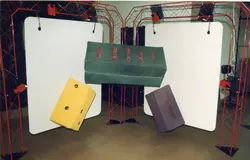 Uma instalação artística ou cenográfica composta por estruturas metálicas vermelhas que sustentam painéis brancos e objetos geométricos suspensos. Os objetos, em tons de verde, amarelo e roxo, parecem ser feitos de material leve ou papelão, sugerindo uma montagem de exposição ou evento cultural.