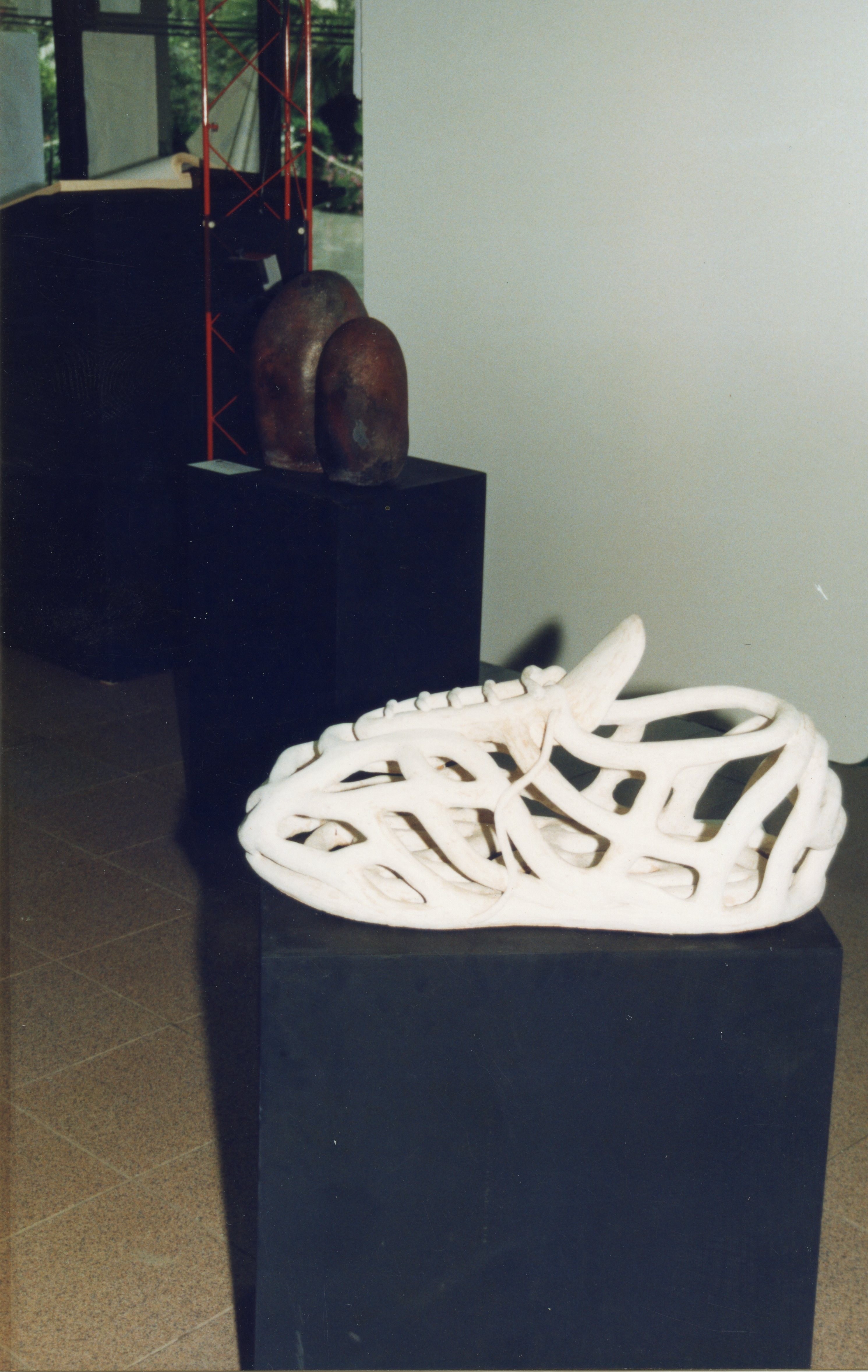 Uma fotografia de uma exposição de arte contemporânea exibindo esculturas sobre pedestais pretos. Em destaque, uma peça branca com estrutura vazada que remete a um calçado, enquanto ao fundo observa-se uma escultura orgânica de tom escuro.