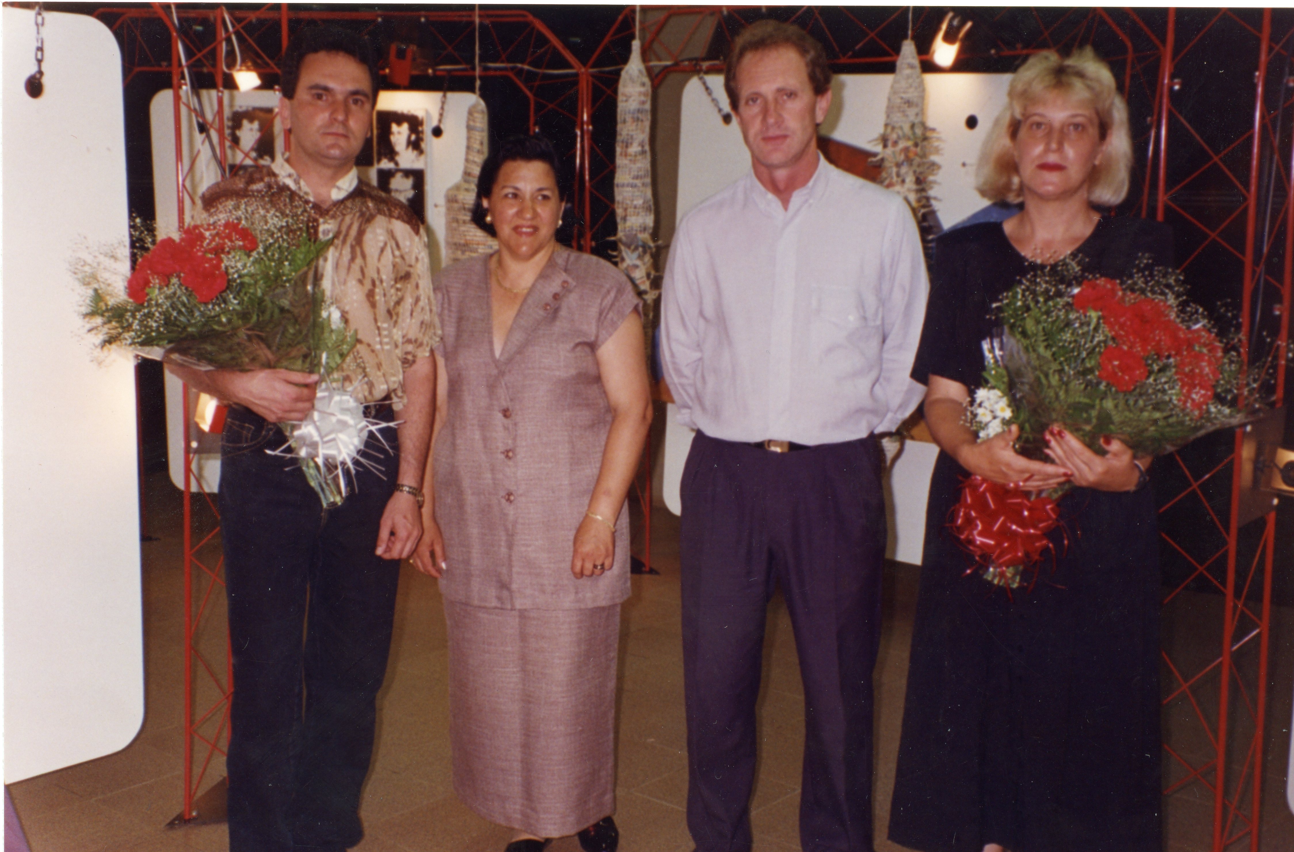 Uma fotografia colorida retratando quatro pessoas posando lado a lado em um ambiente interno, possivelmente uma exposição ou evento cultural. Os indivíduos vestem roupas formais típicas da década de 1990, e dois deles seguram buquês de flores vermelhas, sugerindo uma homenagem ou celebração.
