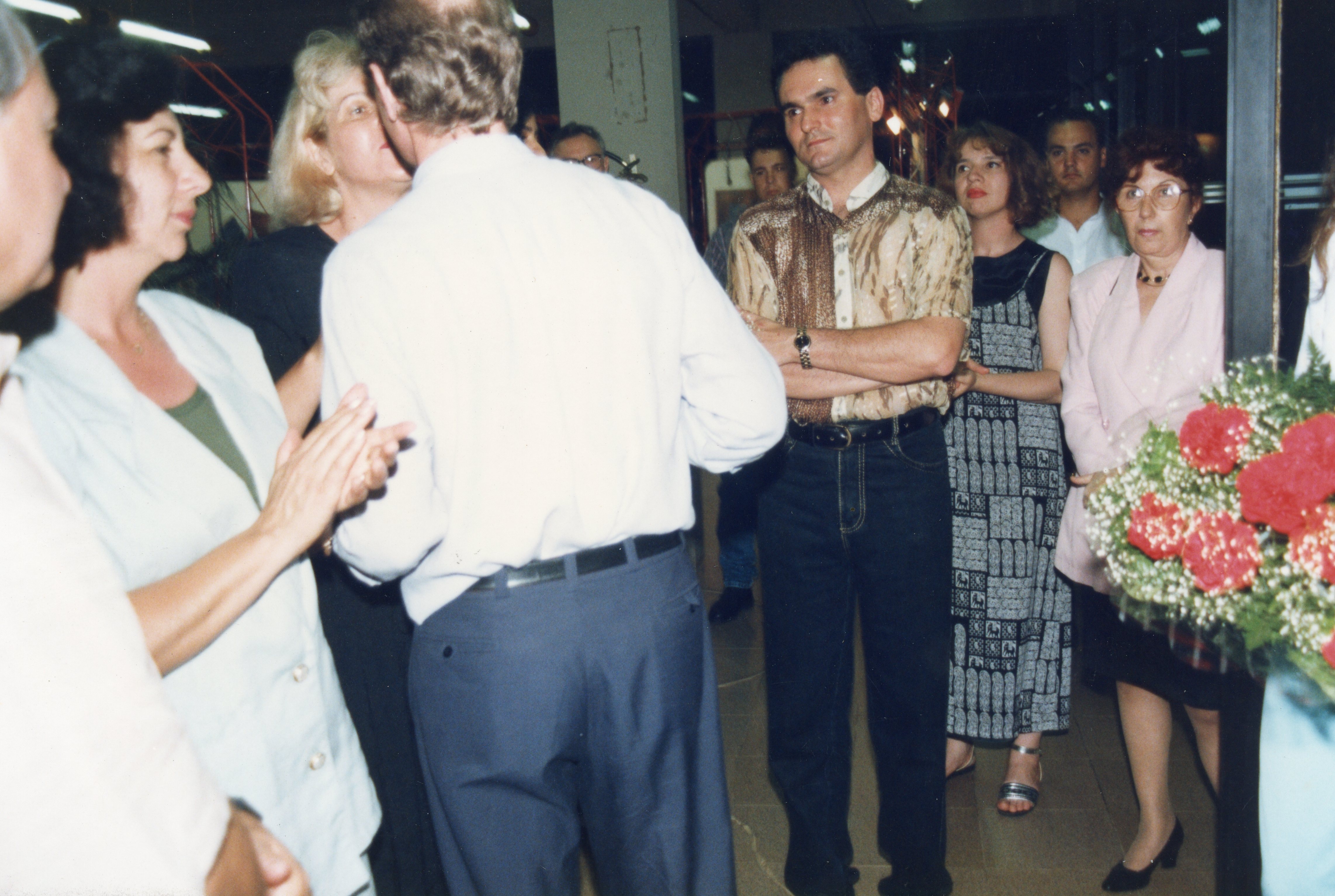 Uma fotografia de um evento social em ambiente interno, possivelmente uma recepção ou exposição, com pessoas vestidas com trajes típicos da década de 1990. O grupo parece estar em um momento de interação, com algumas pessoas aplaudindo e outras observando, destacando-se um homem com camisa estampada e um arranjo de flores em primeiro plano.