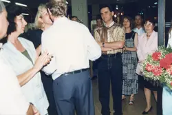 Uma fotografia de um evento social em ambiente interno, possivelmente uma recepção ou exposição, com pessoas vestidas com trajes típicos da década de 1990. O grupo parece estar em um momento de interação, com algumas pessoas aplaudindo e outras observando, destacando-se um homem com camisa estampada e um arranjo de flores em primeiro plano.