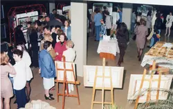 Uma fotografia de um evento social, possivelmente uma exposição de arte ou vernissage, realizada em um ambiente interno. O público, composto por homens e mulheres com vestimentas típicas da década de 1990, circula entre cavaletes com quadros e mesas com alimentos, sugerindo um contexto de confraternização cultural em uma cidade brasileira.