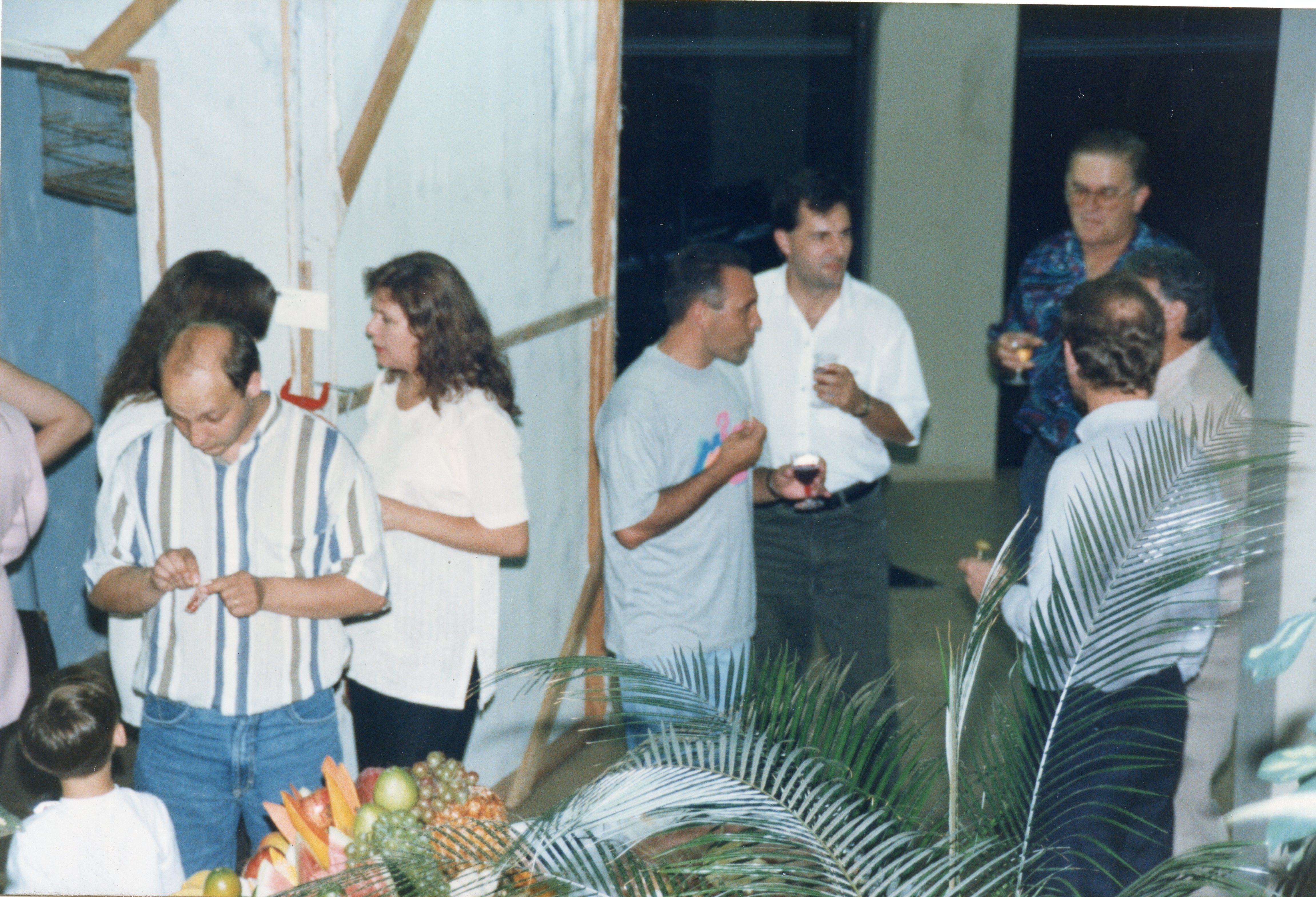 Uma fotografia de um evento social em ambiente interno, possivelmente uma confraternização ou recepção, com um grupo de adultos conversando e consumindo bebidas. As vestimentas, incluindo camisas polo e camisetas estampadas, sugerem um contexto casual típico das décadas de 1980 ou 1990.
