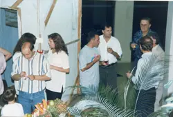 Uma fotografia de um evento social em ambiente interno, possivelmente uma confraternização ou recepção, com um grupo de adultos conversando e consumindo bebidas. As vestimentas, incluindo camisas polo e camisetas estampadas, sugerem um contexto casual típico das décadas de 1980 ou 1990.