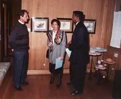 Uma fotografia de época retratando três pessoas em uma galeria de arte ou exposição cultural. O ambiente possui paredes e piso de madeira, com quadros pendurados e esculturas decorativas, sugerindo um evento social ou vernissage ocorrido provavelmente na década de 1990.