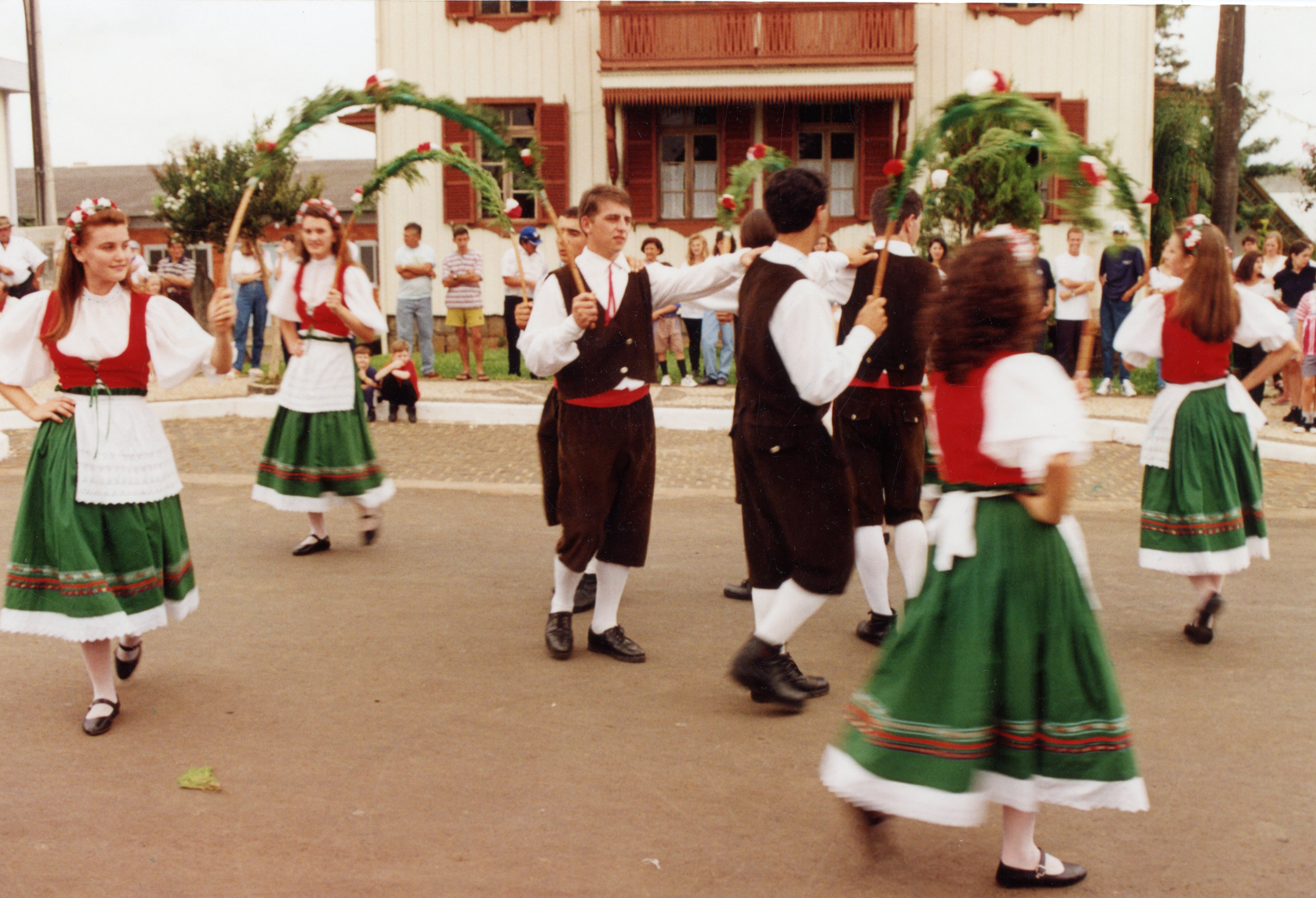 Um grupo de jovens dançarinos apresenta uma dança folclórica tradicional, possivelmente de origem europeia, em frente a uma construção de madeira com arquitetura típica de colonização. Os participantes vestem trajes tradicionais com saias verdes, aventais brancos e coletes, segurando arcos decorados com flores, sugerindo um evento cultural ou festividade comunitária.