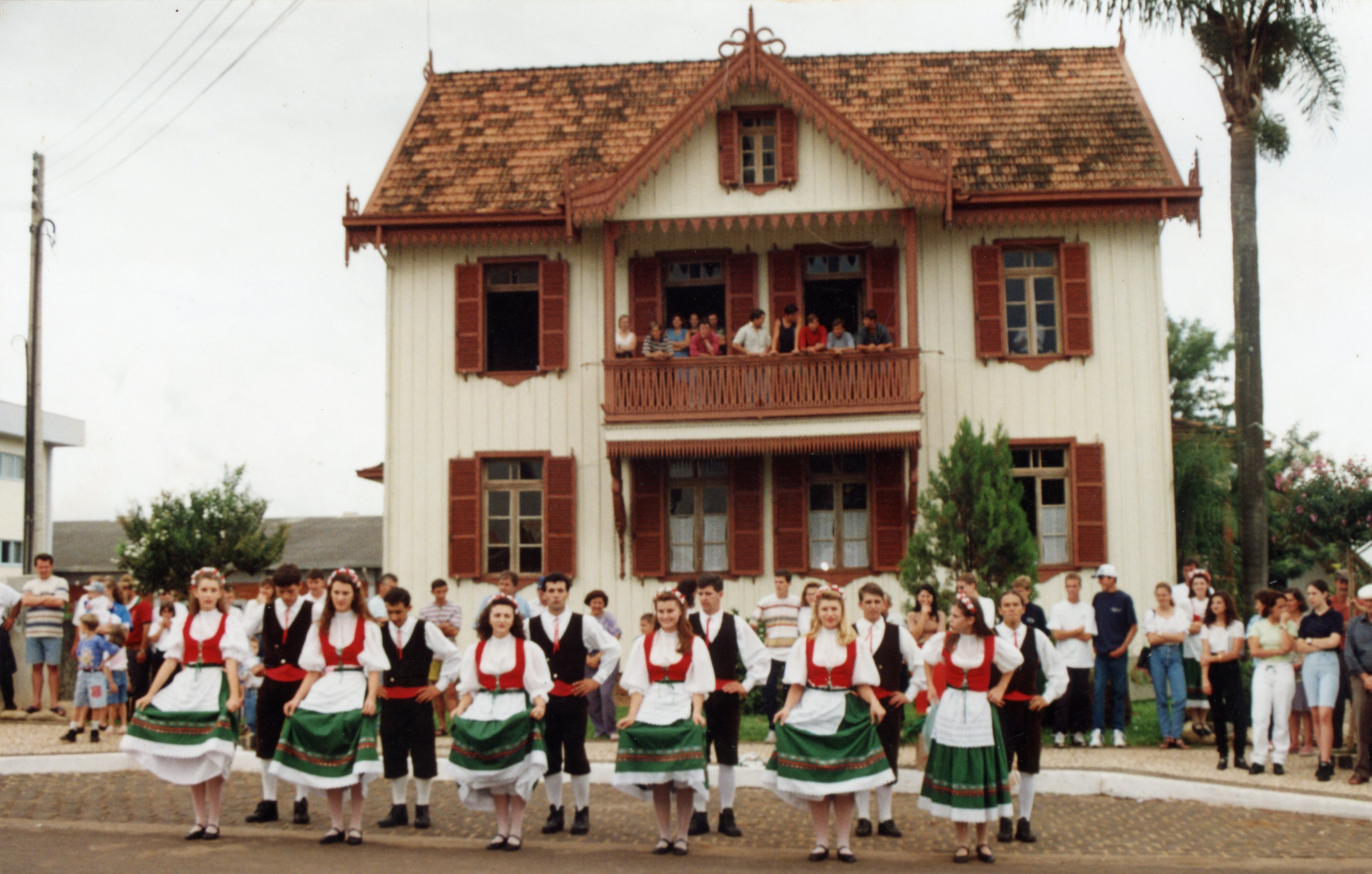 Uma fotografia colorida que registra um grupo de dançarinos folclóricos em trajes típicos europeus, possivelmente de origem italiana ou alemã, apresentando-se em frente a uma casa de madeira de arquitetura tradicional. A cena sugere um evento cultural ou festividade comunitária, com espectadores observando tanto do térreo quanto da sacada do casarão.