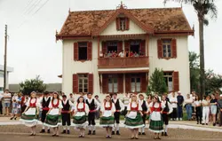 Uma fotografia colorida que registra um grupo de dançarinos folclóricos em trajes típicos europeus, possivelmente de origem italiana ou alemã, apresentando-se em frente a uma casa de madeira de arquitetura tradicional. A cena sugere um evento cultural ou festividade comunitária, com espectadores observando tanto do térreo quanto da sacada do casarão.