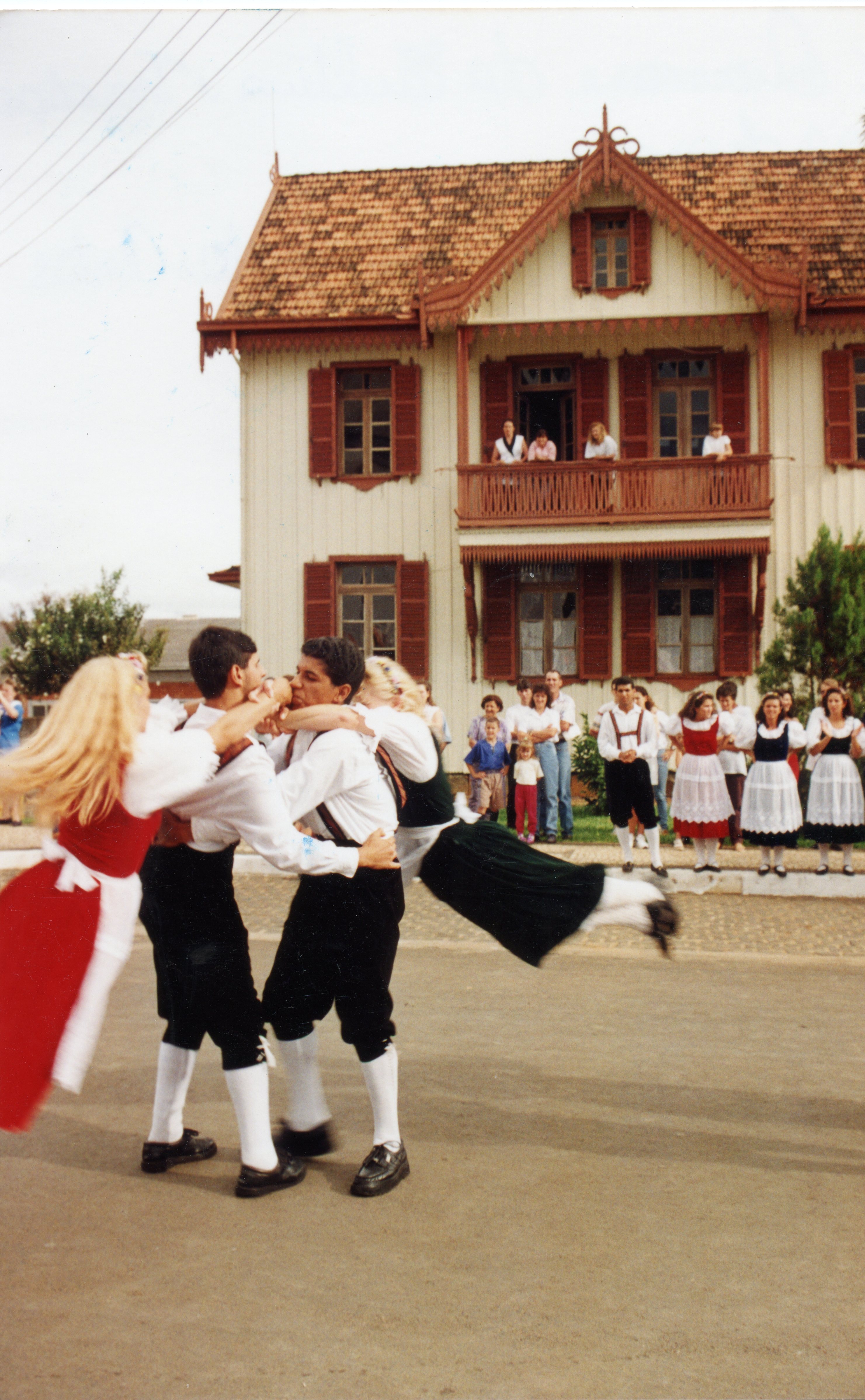 Uma fotografia colorida capturando um grupo de jovens realizando uma dança folclórica tradicional em frente a uma casa de arquitetura estilo enxaimel. O cenário sugere uma celebração cultural, possivelmente em uma comunidade de descendência europeia no sul do Brasil, com os participantes vestindo trajes típicos.