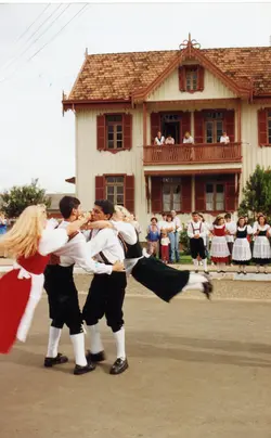 Uma fotografia colorida capturando um grupo de jovens realizando uma dança folclórica tradicional em frente a uma casa de arquitetura estilo enxaimel. O cenário sugere uma celebração cultural, possivelmente em uma comunidade de descendência europeia no sul do Brasil, com os participantes vestindo trajes típicos.
