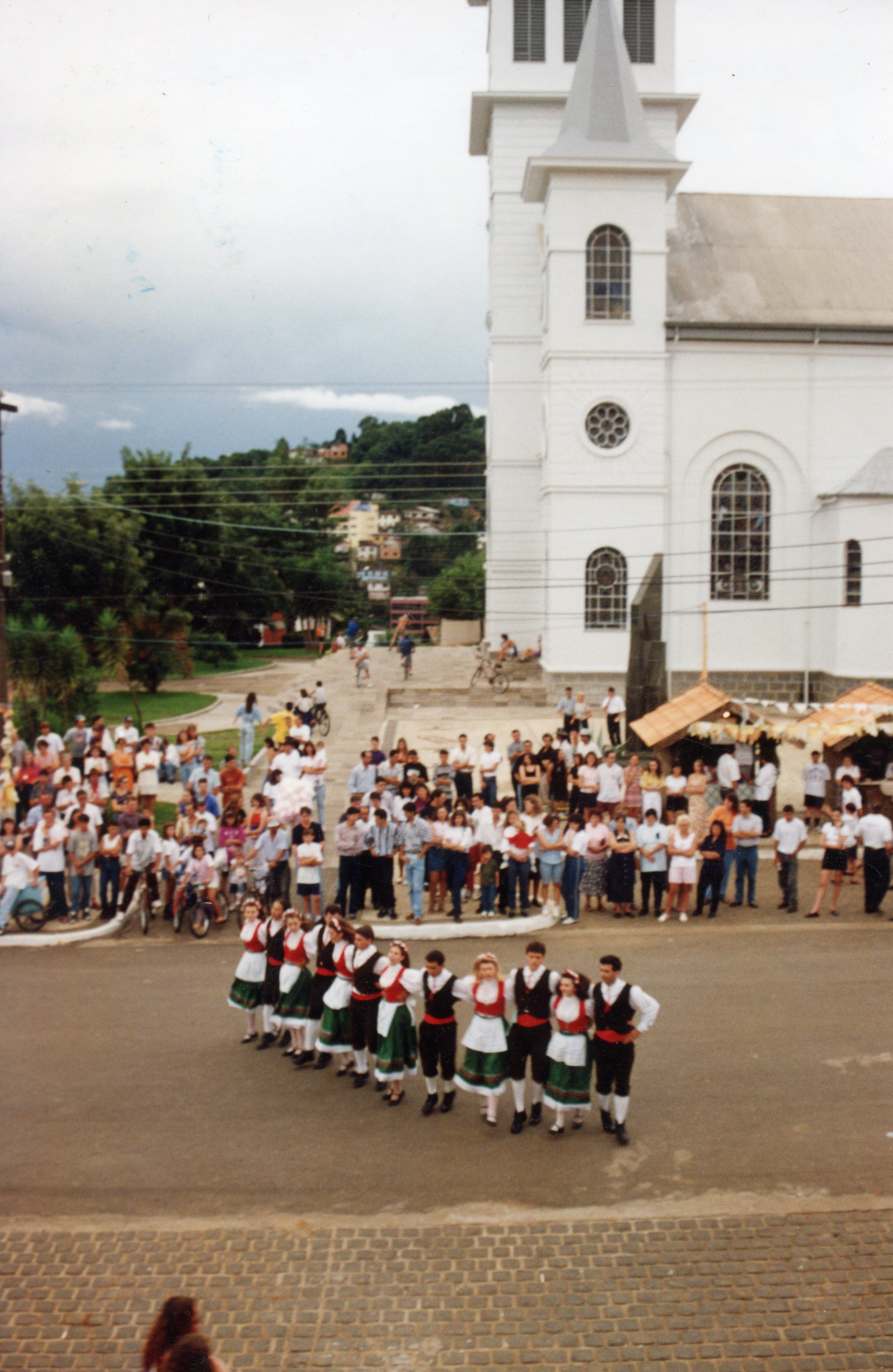 Uma fotografia colorida que captura um grupo de dançarinos folclóricos em trajes tradicionais europeus, possivelmente de origem italiana ou alemã, apresentando-se em uma praça pública. Ao fundo, observa-se uma igreja de arquitetura clássica e uma multidão de espectadores, sugerindo um evento cultural ou festividade comunitária em uma cidade do interior do Brasil, possivelmente nas décadas de 1990 ou 2000.