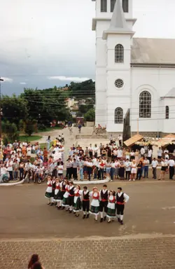 Uma fotografia colorida que captura um grupo de dançarinos folclóricos em trajes tradicionais europeus, possivelmente de origem italiana ou alemã, apresentando-se em uma praça pública. Ao fundo, observa-se uma igreja de arquitetura clássica e uma multidão de espectadores, sugerindo um evento cultural ou festividade comunitária em uma cidade do interior do Brasil, possivelmente nas décadas de 1990 ou 2000.
