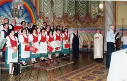 Uma fotografia colorida que registra um coral em apresentação dentro de uma igreja, possivelmente em um contexto de celebração comunitária ou religiosa. Os integrantes vestem trajes típicos, sugerindo uma influência cultural europeia, possivelmente italiana, comum em cidades como Videira, com um homem discursando ao microfone à direita.