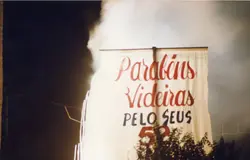 Uma fotografia noturna exibindo um grande cartaz comemorativo com a inscrição 'Parabéns Videiras PELO SEUS 52'. O elemento sugere uma celebração de aniversário da cidade de Videira, possivelmente realizada em um espaço público ou evento festivo.