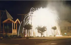 Uma fotografia noturna capturando uma exibição de fogos de artifício, possivelmente uma cascata, em uma praça ou área pública. A cena mostra uma estrutura de madeira com telhado decorado à esquerda e casas ao fundo, sugerindo um ambiente urbano de uma pequena cidade brasileira.
