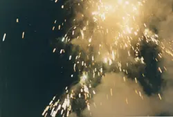 Uma fotografia noturna capturando o rastro luminoso e a explosão de fogos de artifício no céu. A imagem apresenta um alto contraste entre a escuridão do ambiente e o brilho intenso das faíscas, sugerindo um evento comemorativo ou festivo.