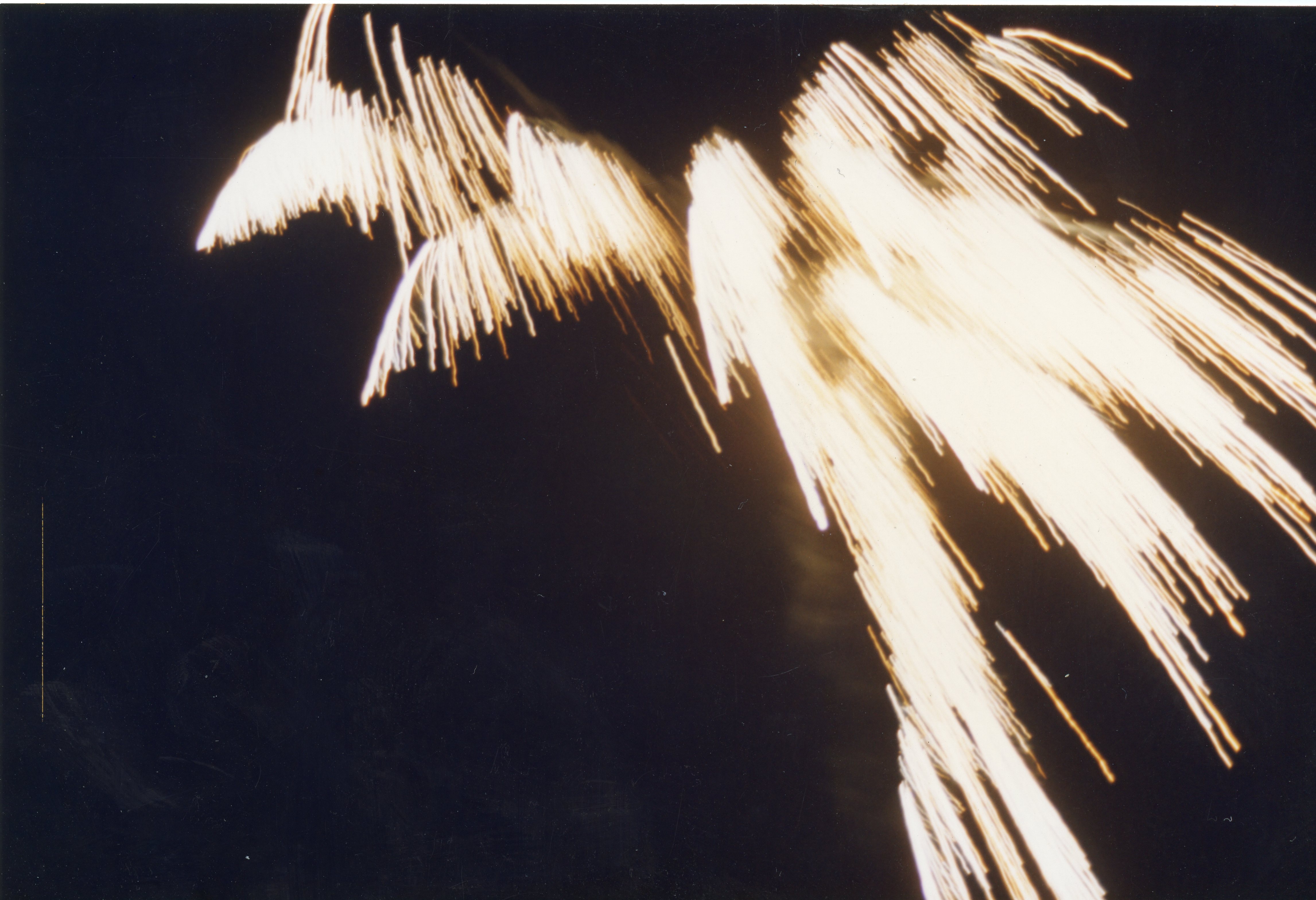 Uma fotografia de longa exposição capturando o rastro luminoso de fogos de artifício contra um céu noturno escuro. A imagem apresenta linhas de luz brilhantes e dinâmicas, sugerindo um evento comemorativo ou festivo.