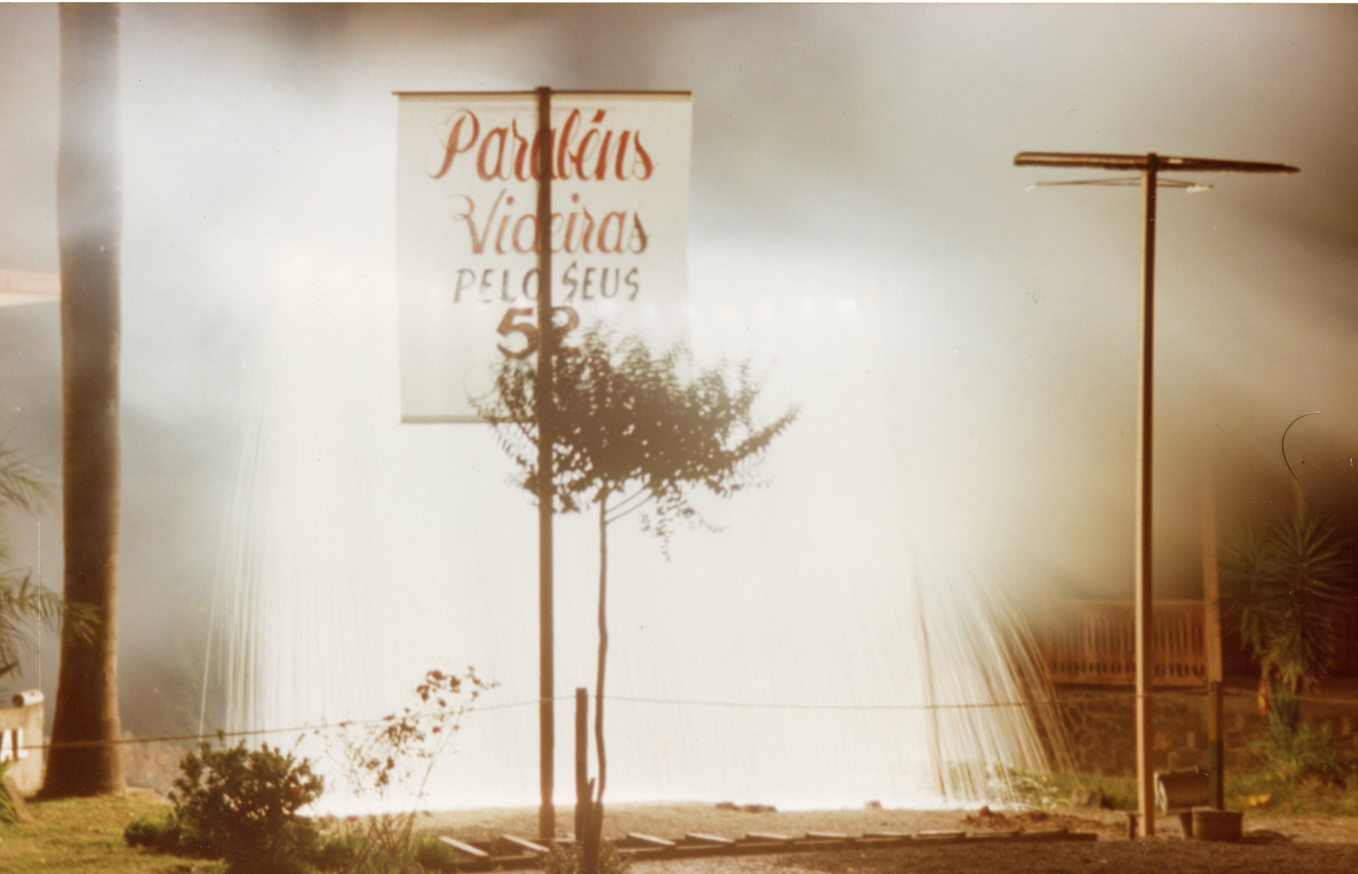 Uma fotografia noturna que exibe uma placa comemorativa com os dizeres 'Parabéns Videira pelo seus 52', indicando uma celebração de aniversário da cidade. A imagem apresenta um efeito de luz estourada ou neblina, sugerindo uma captura feita com flash ou iluminação intensa em um ambiente externo, possivelmente em uma praça ou via pública.